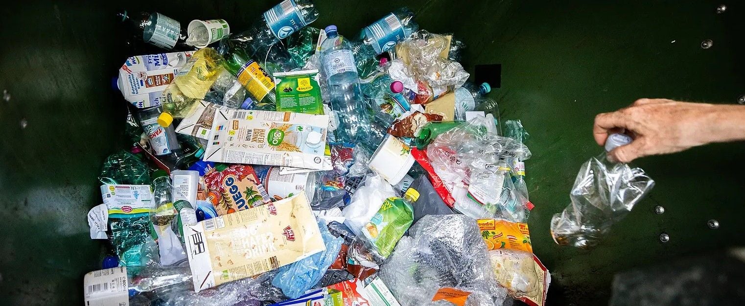 Verschiedene Plastikabfälle und Verpackungen in einem Müllcontainer, eine Hand wirft Müll hinein.