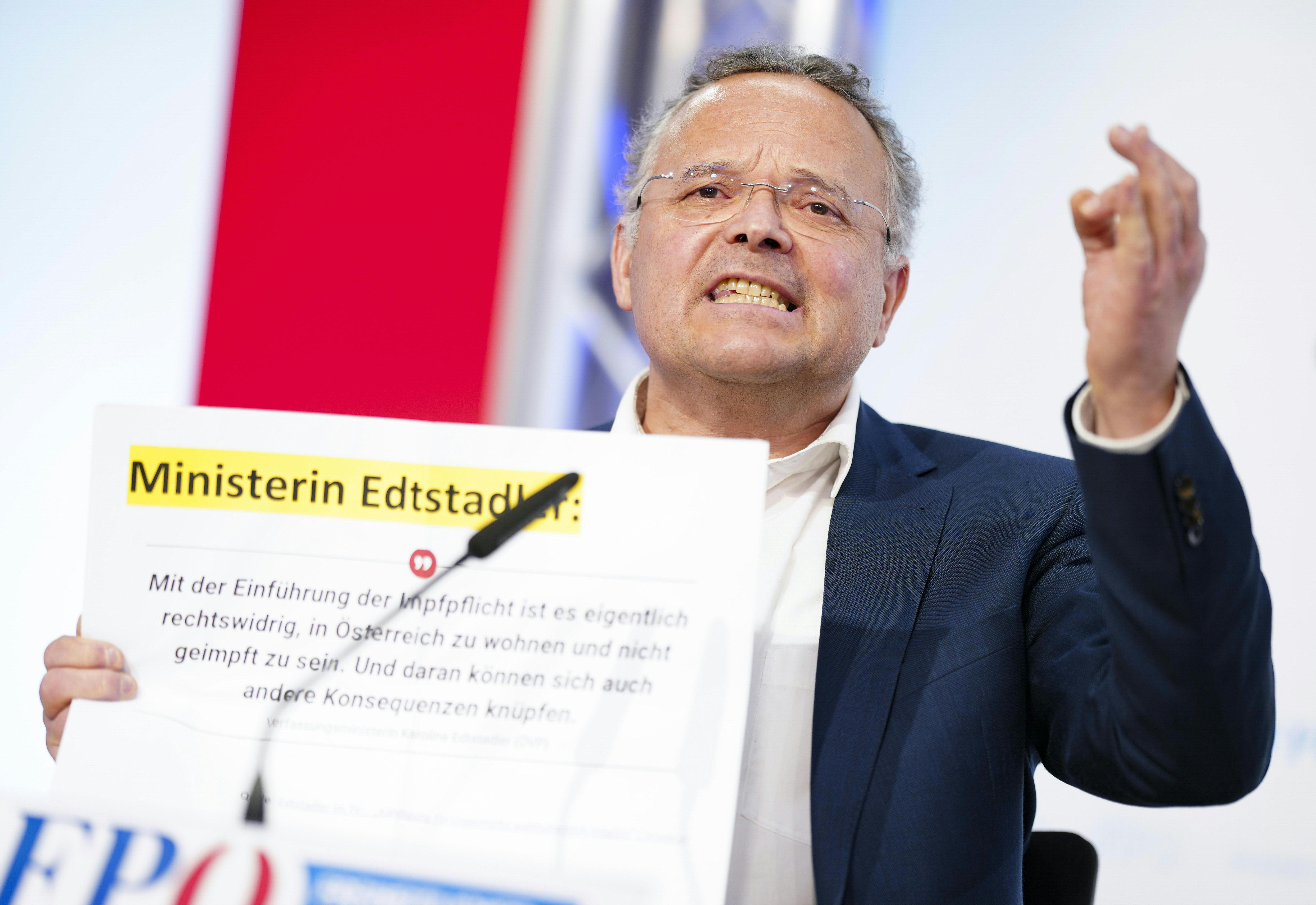 Ein Mann im Anzug hält ein Schild mit Text während einer Pressekonferenz hoch.