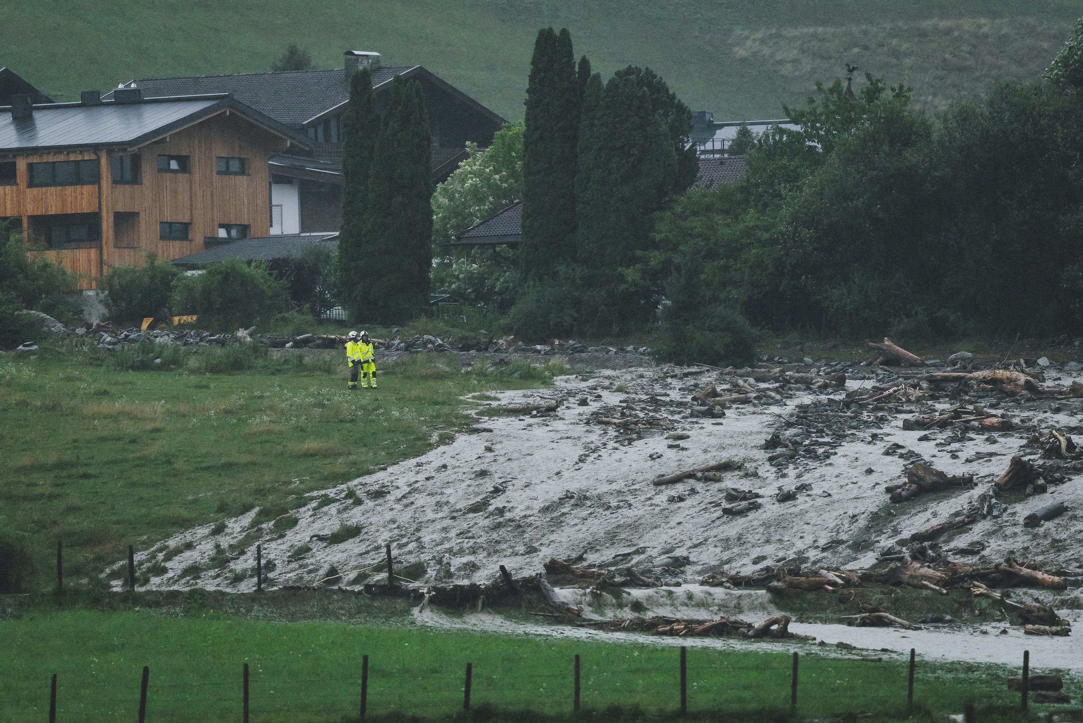 Zwei Einsatzkräfte begutachten eine überschwemmte und vermurte Wiese nahe Häusern in Uttendorf.