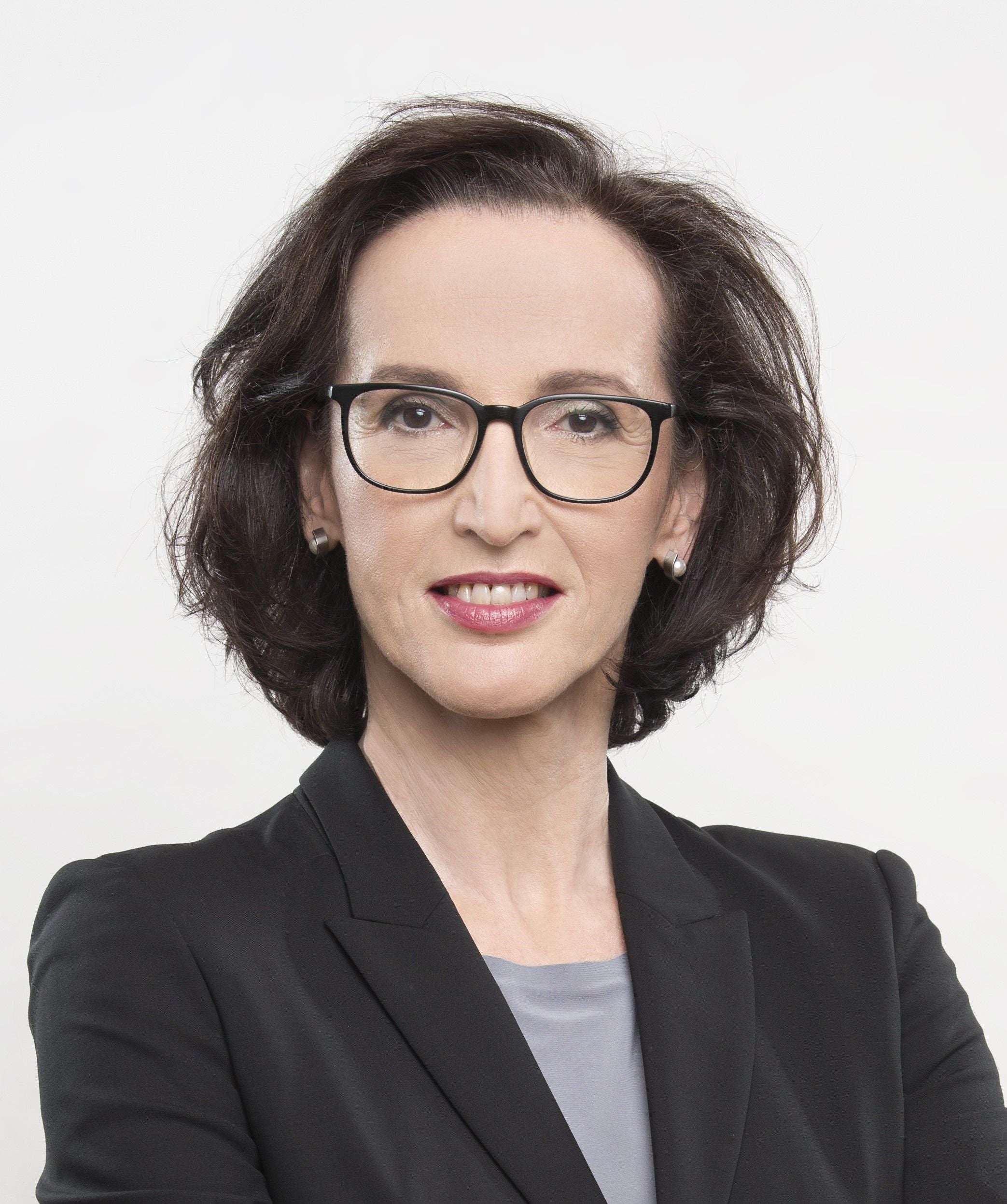 Frau mit dunklen Haaren, Brille und dunklem Blazer vor weißem Hintergrund.