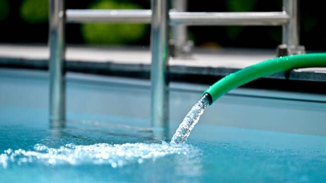 Ein grüner Gartenschlauch füllt Wasser in einen Pool neben einer Metallleiter.
