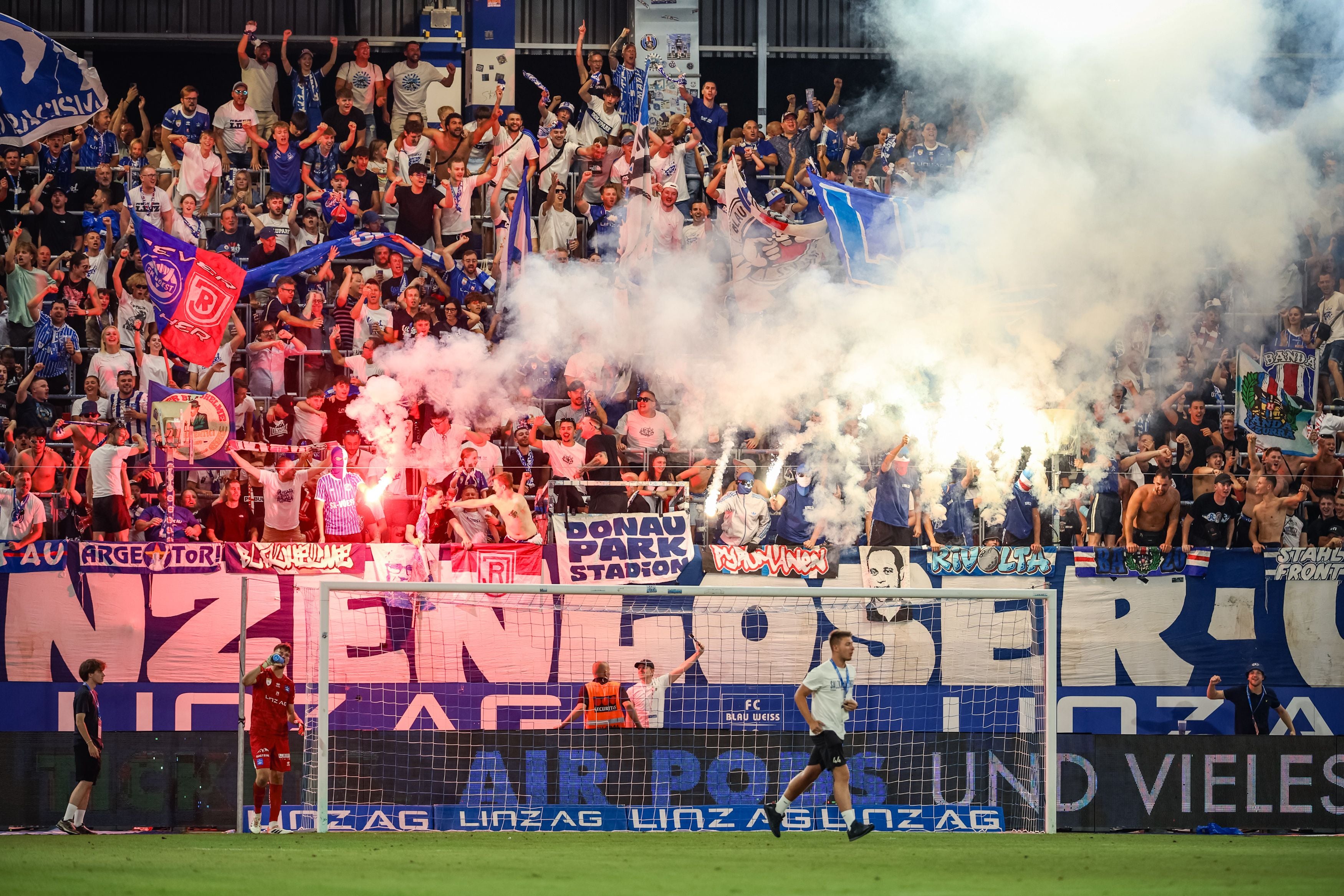 Fußballfans von Blau-Weiß Linz jubeln im Stadion mit Pyrotechnik und Fahnen hinter dem Tor.
