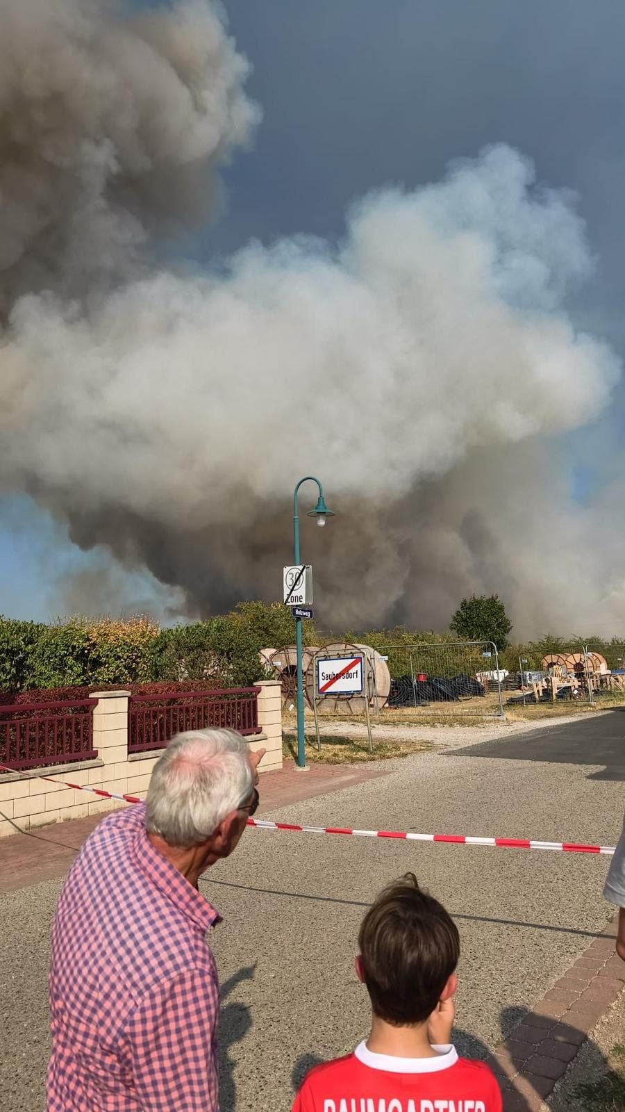 Dichter Rauch steigt bei einem Waldbrand auf, Menschen beobachten das Geschehen hinter Absperrband.