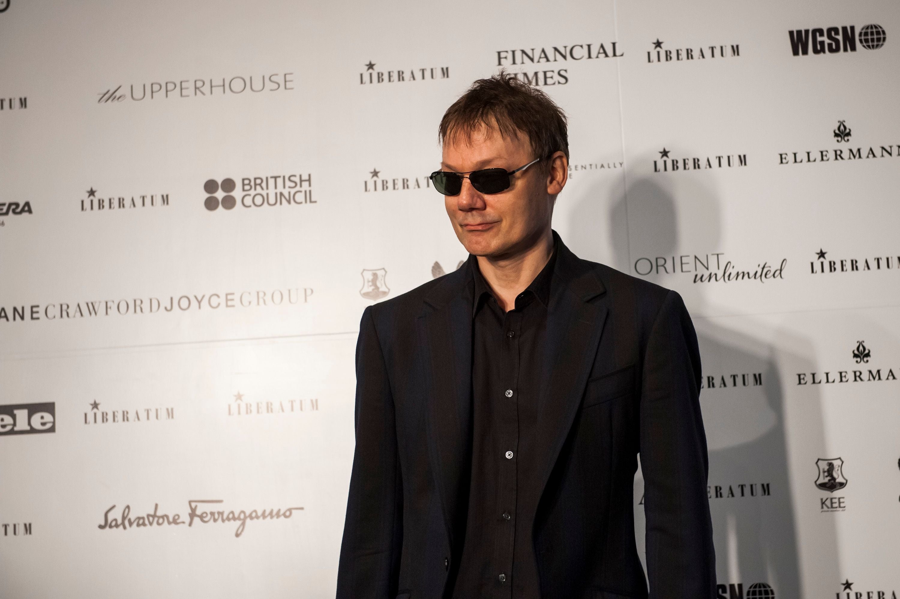 Mann mit Sonnenbrille im schwarzen Anzug auf einem Event mit Sponsorenwand im Hintergrund.