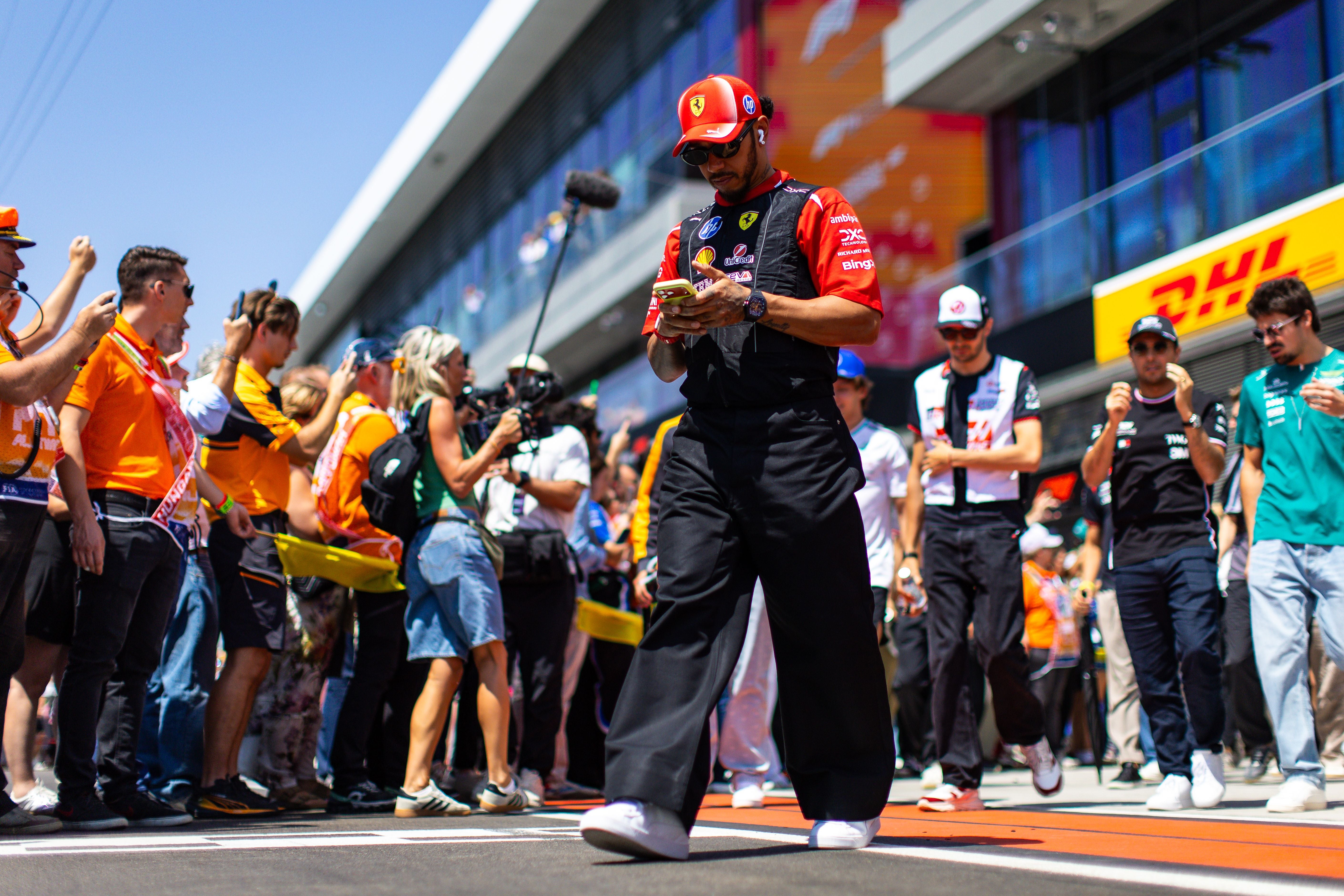 Ein Formel-1-Fahrer im Ferrari-Outfit läuft mit Handy durch das Fahrerlager, umgeben von Fans und Teams.