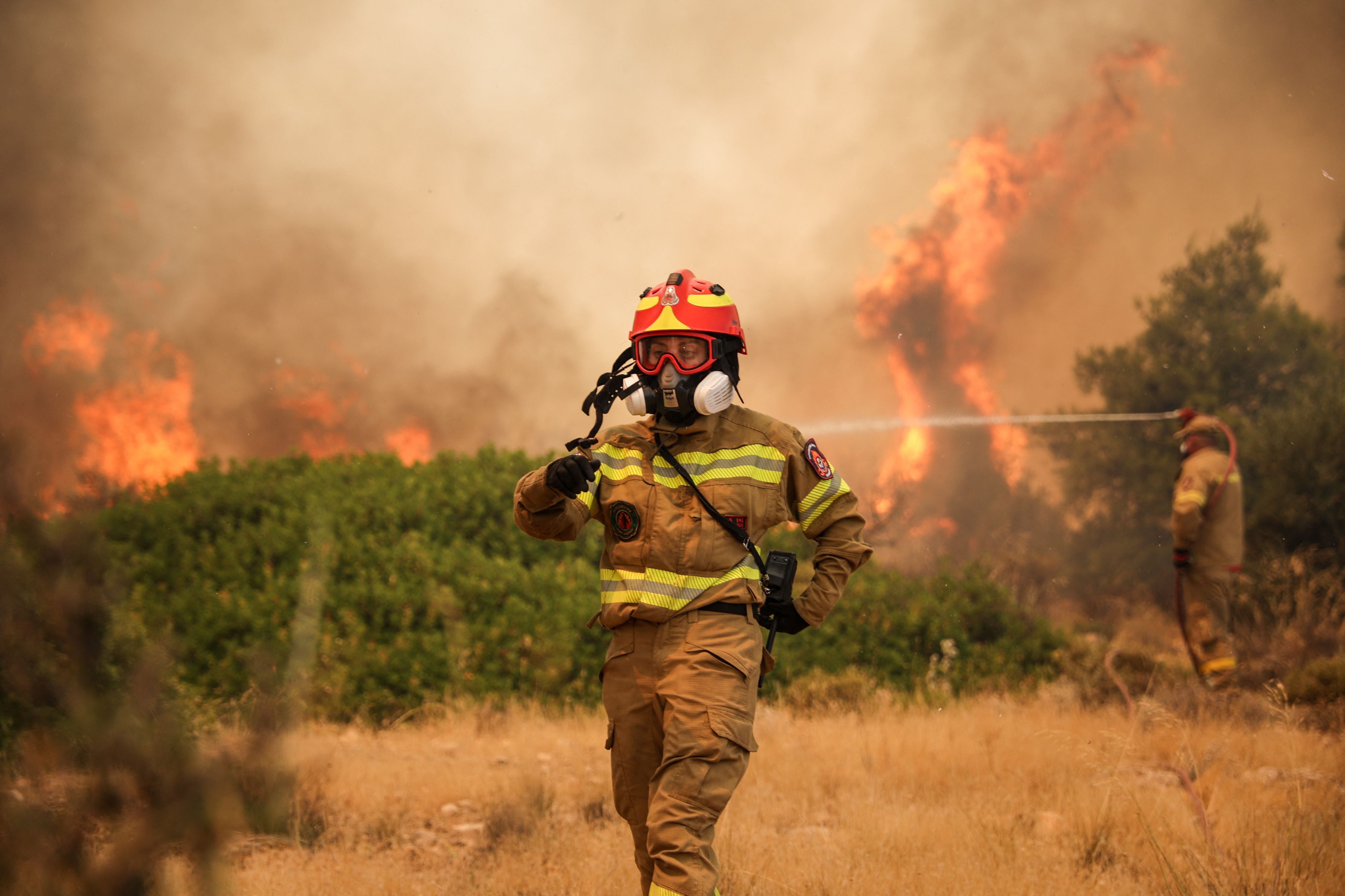 Feuerwehrleute bekämpfen einen Waldbrand in einer trockenen, bewachsenen Landschaft bei starker Rauchentwicklung.
