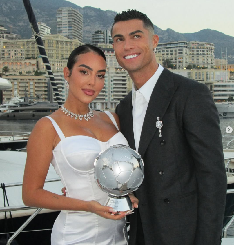 Eine Frau in weißem Kleid und ein Mann im Anzug stehen mit einer Trophäe vor Yachten und Gebäuden.