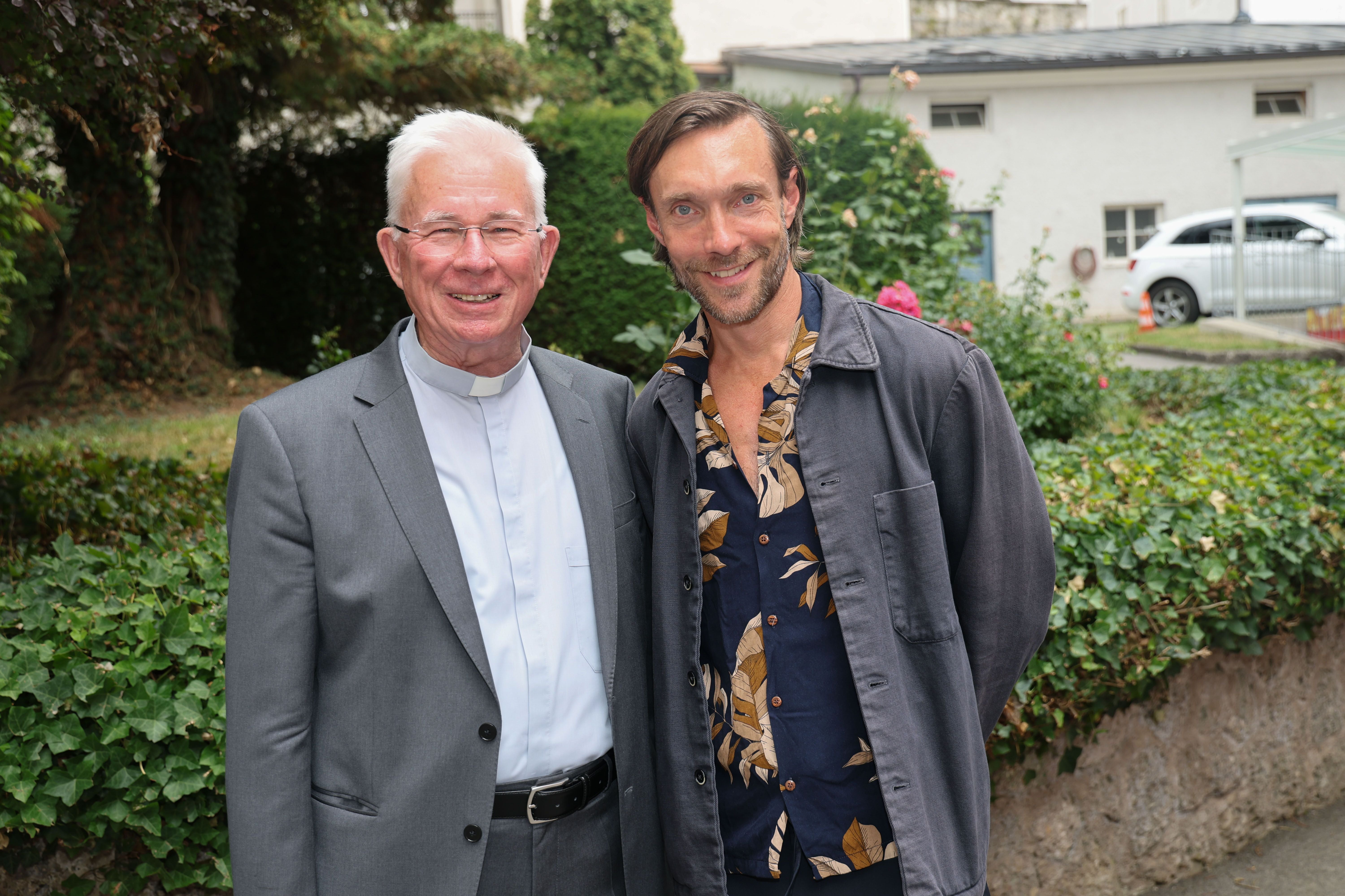 Zwei Männer lächeln im Freien in die Kamera, einer trägt Priesterkleidung, der andere Freizeitkleidung.