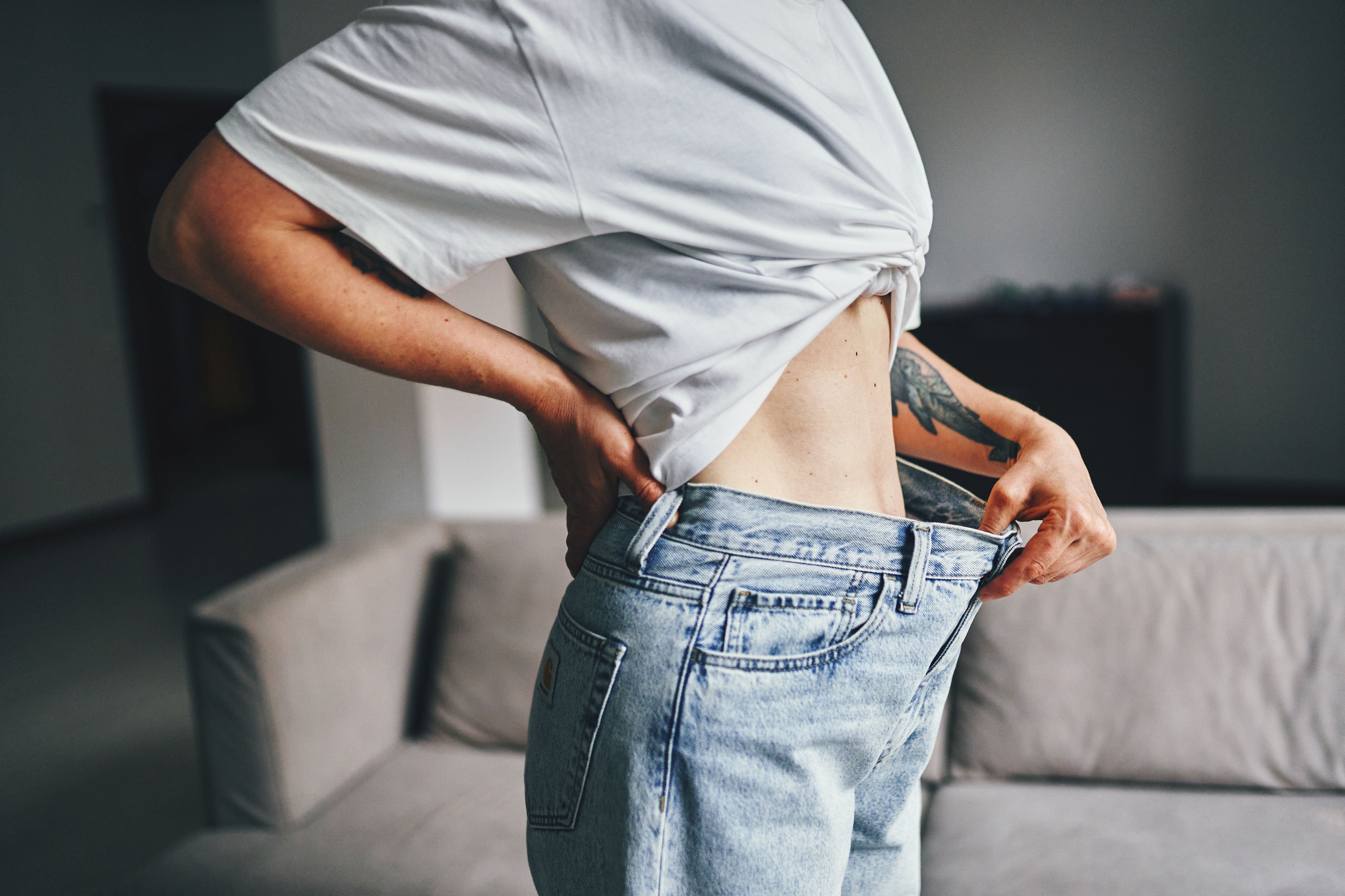 Person zeigt Gewichtsverlust, indem sie lose Jeans in einem Wohnzimmer trägt.