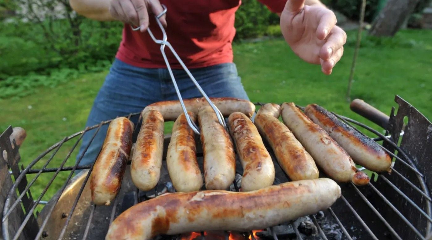 Mehrere Bratwürste liegen auf einem Grill; eine Person wendet sie mit einer Grillzange.