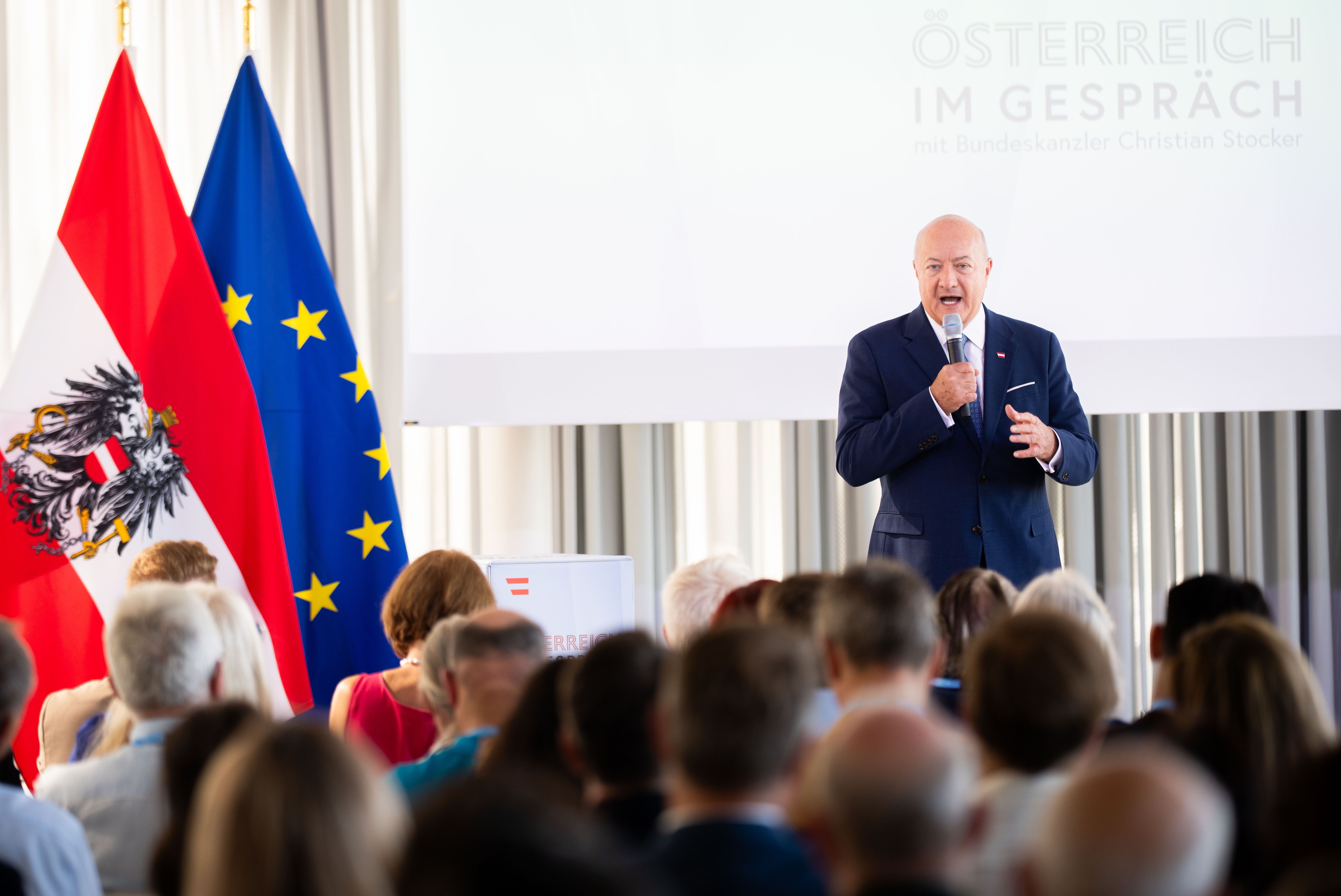 Ein Mann spricht bei einer Veranstaltung vor Publikum, daneben Österreich- und EU-Flagge.