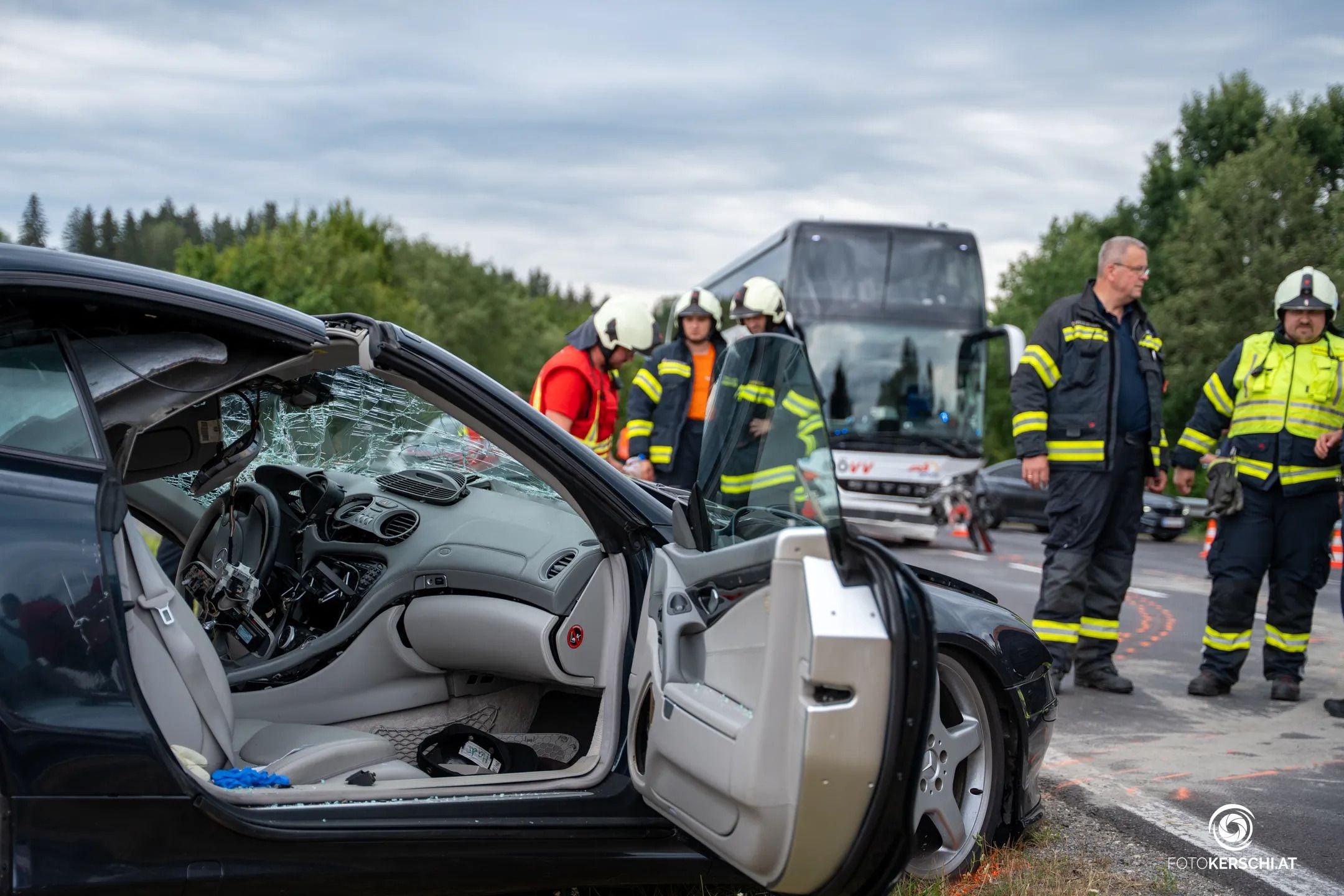Verunfalltes Auto mit eingeschlagener Frontscheibe, Feuerwehrleute und Bus im Hintergrund.