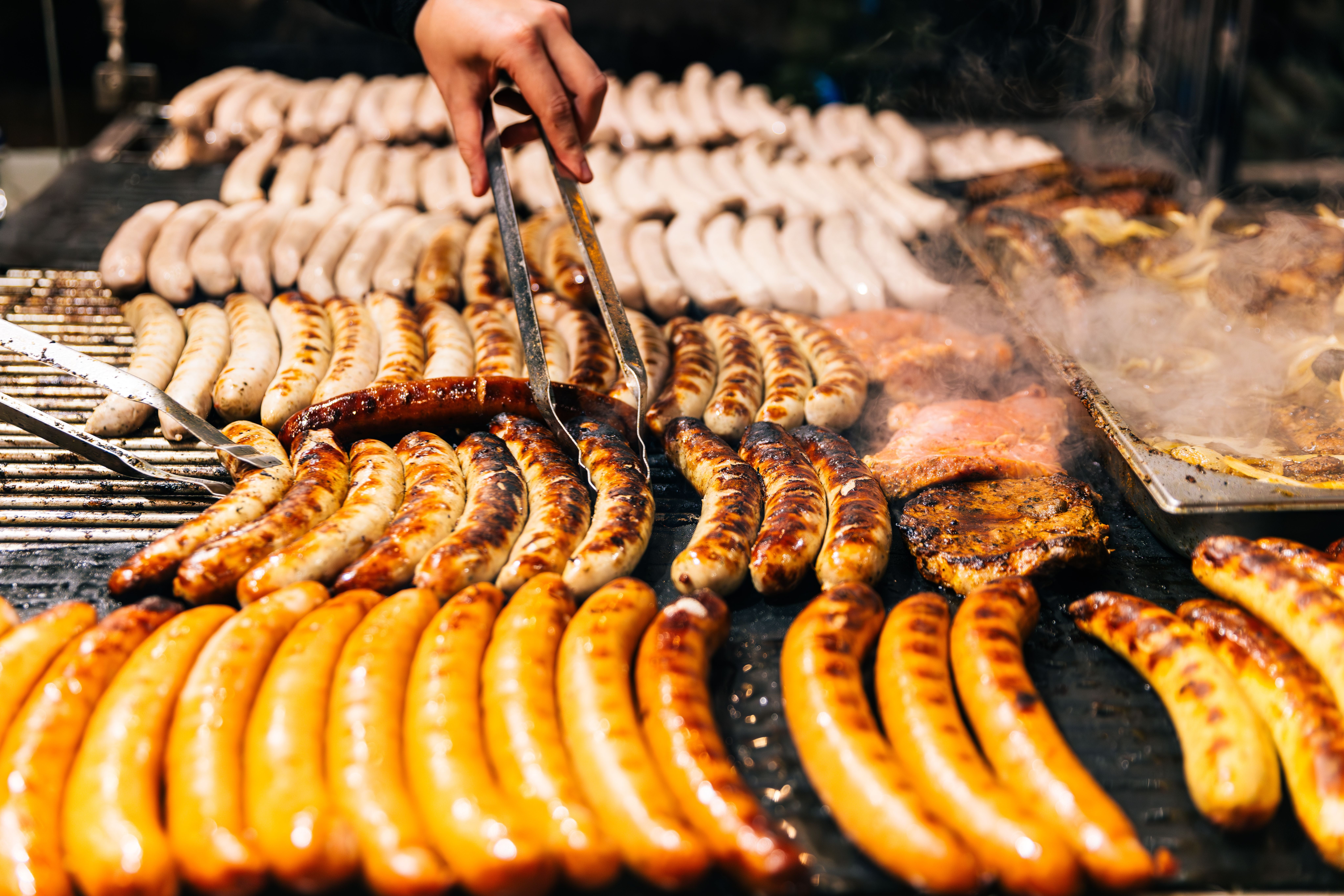 Viele Bratwürste und Fleischstücke werden auf einem Grill mit einer Zange gewendet.