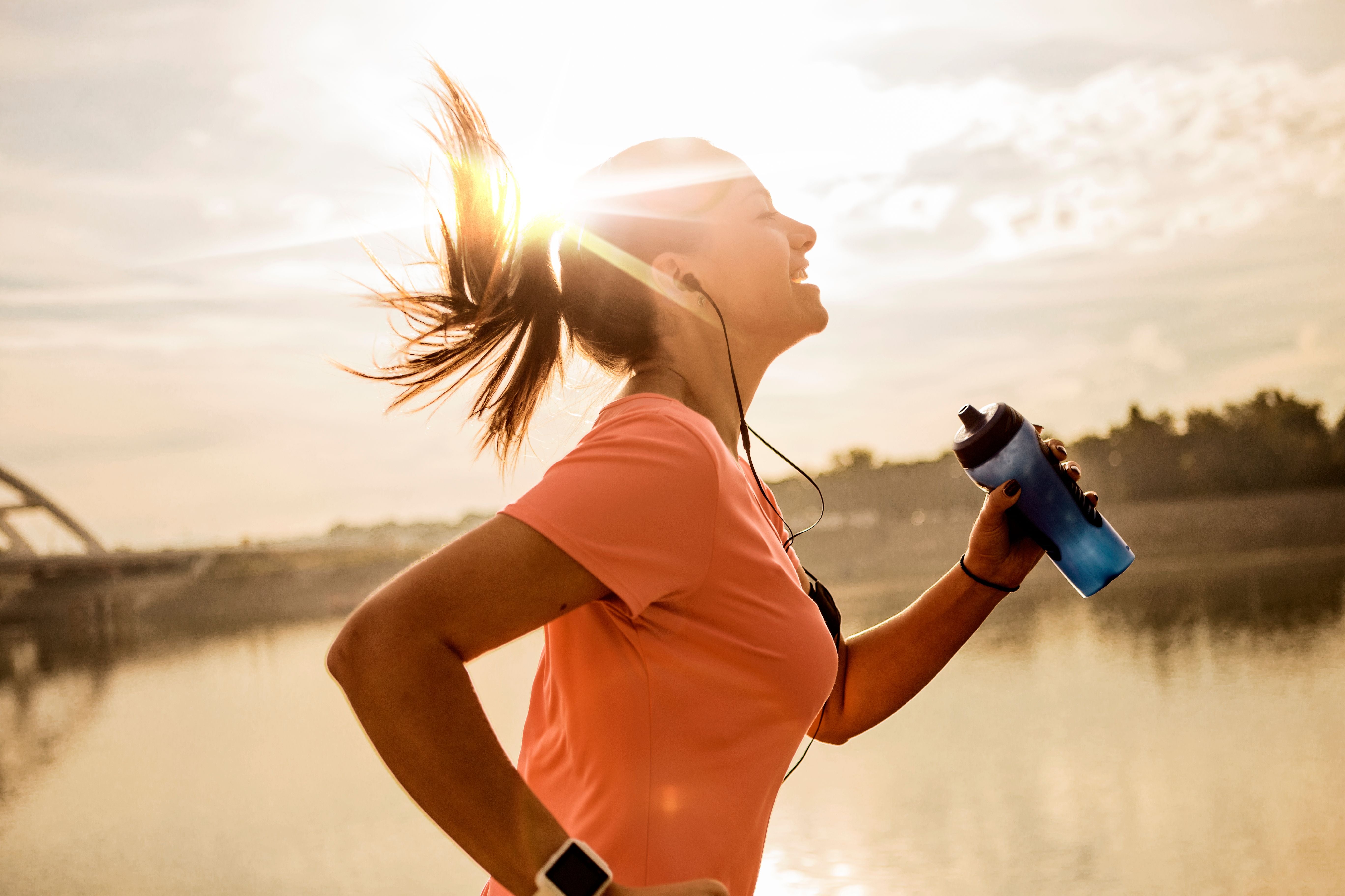 Junge Frau joggt am Wasser, hält eine Trinkflasche und lächelt in der Sonne.