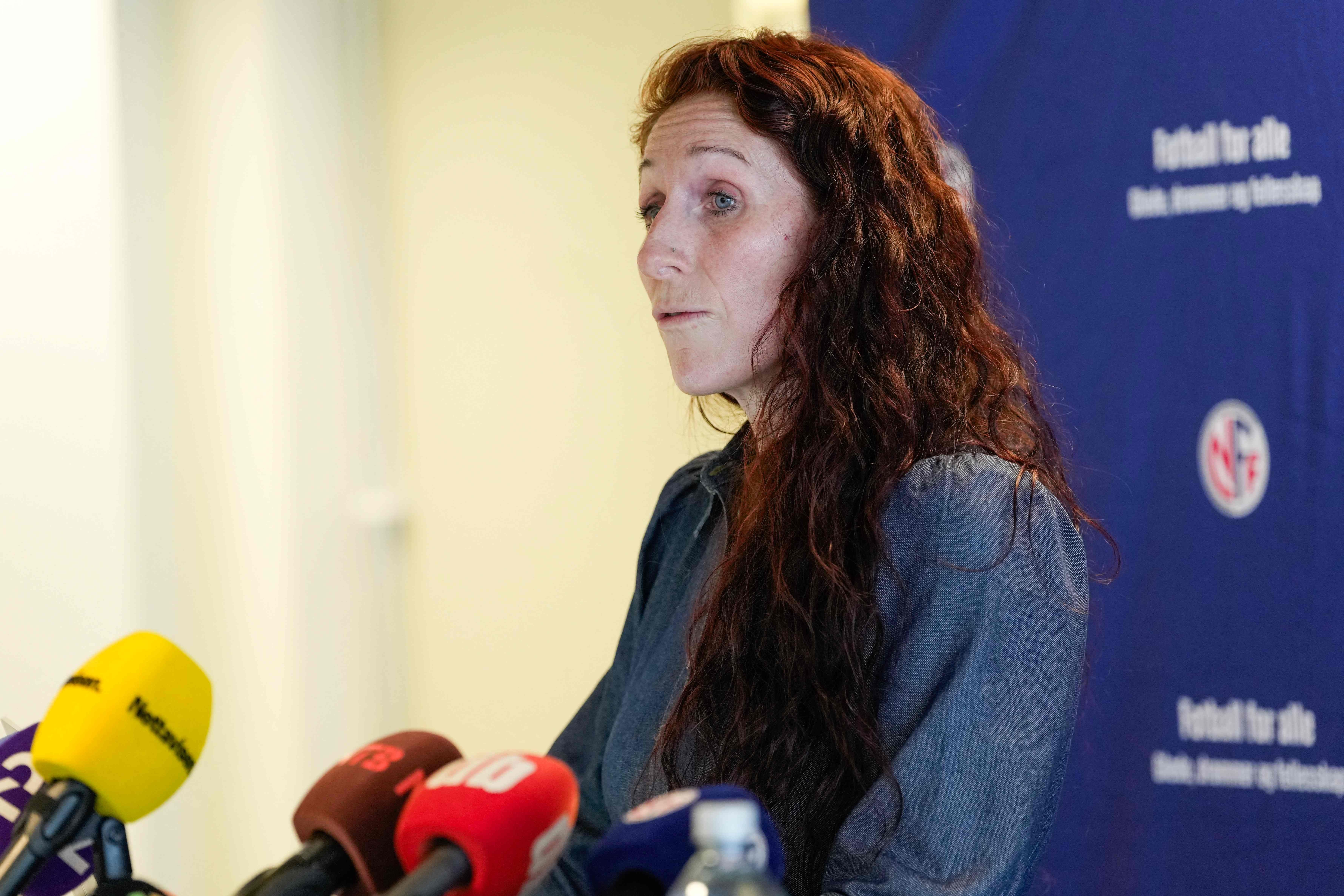 Eine Frau mit langen, lockigen Haaren spricht auf einer Pressekonferenz vor mehreren Mikrofonen.