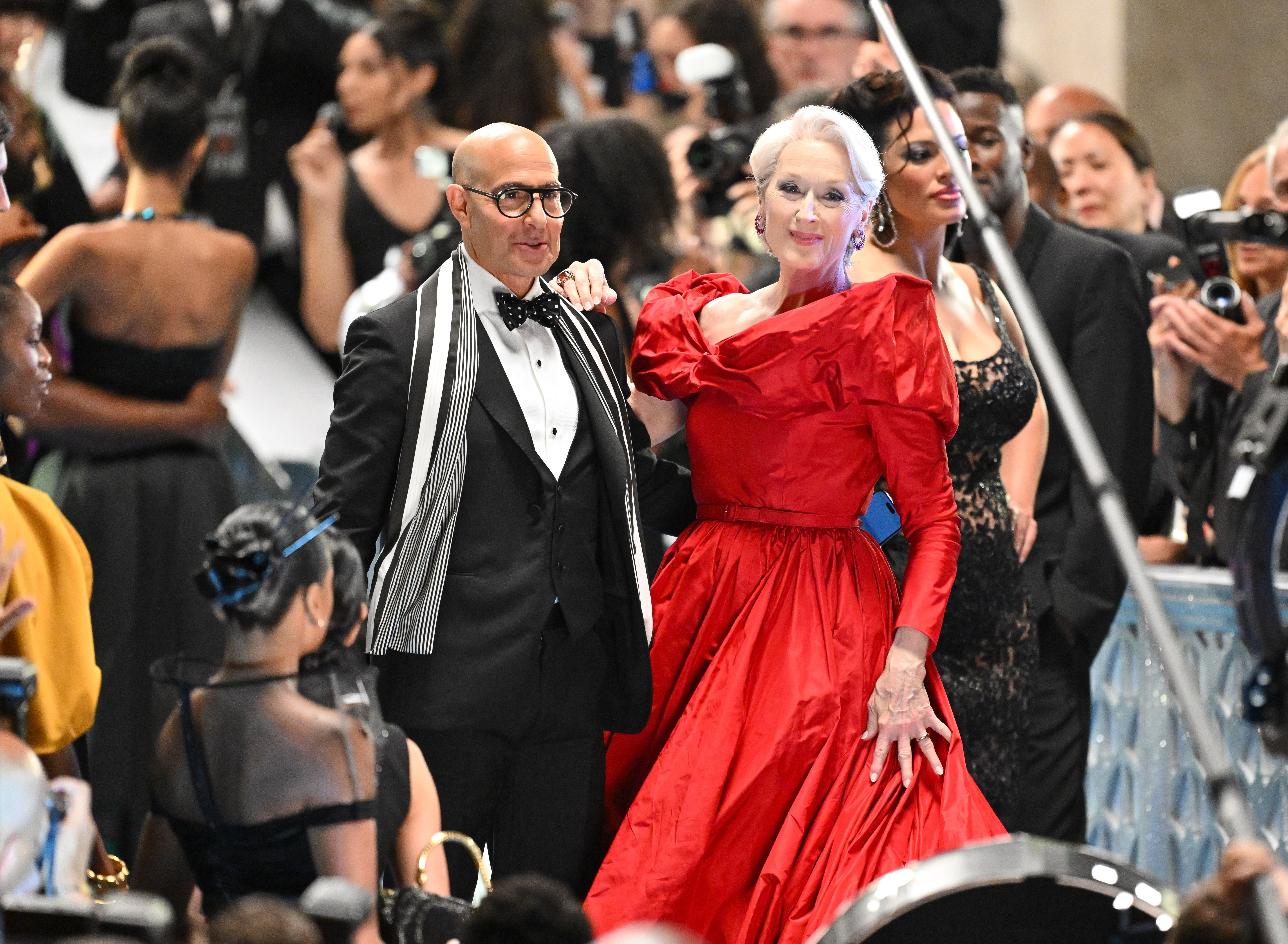 Ein Mann im Smoking und eine Frau im roten Kleid stehen auf einem eleganten Event, umgeben von Menschen.