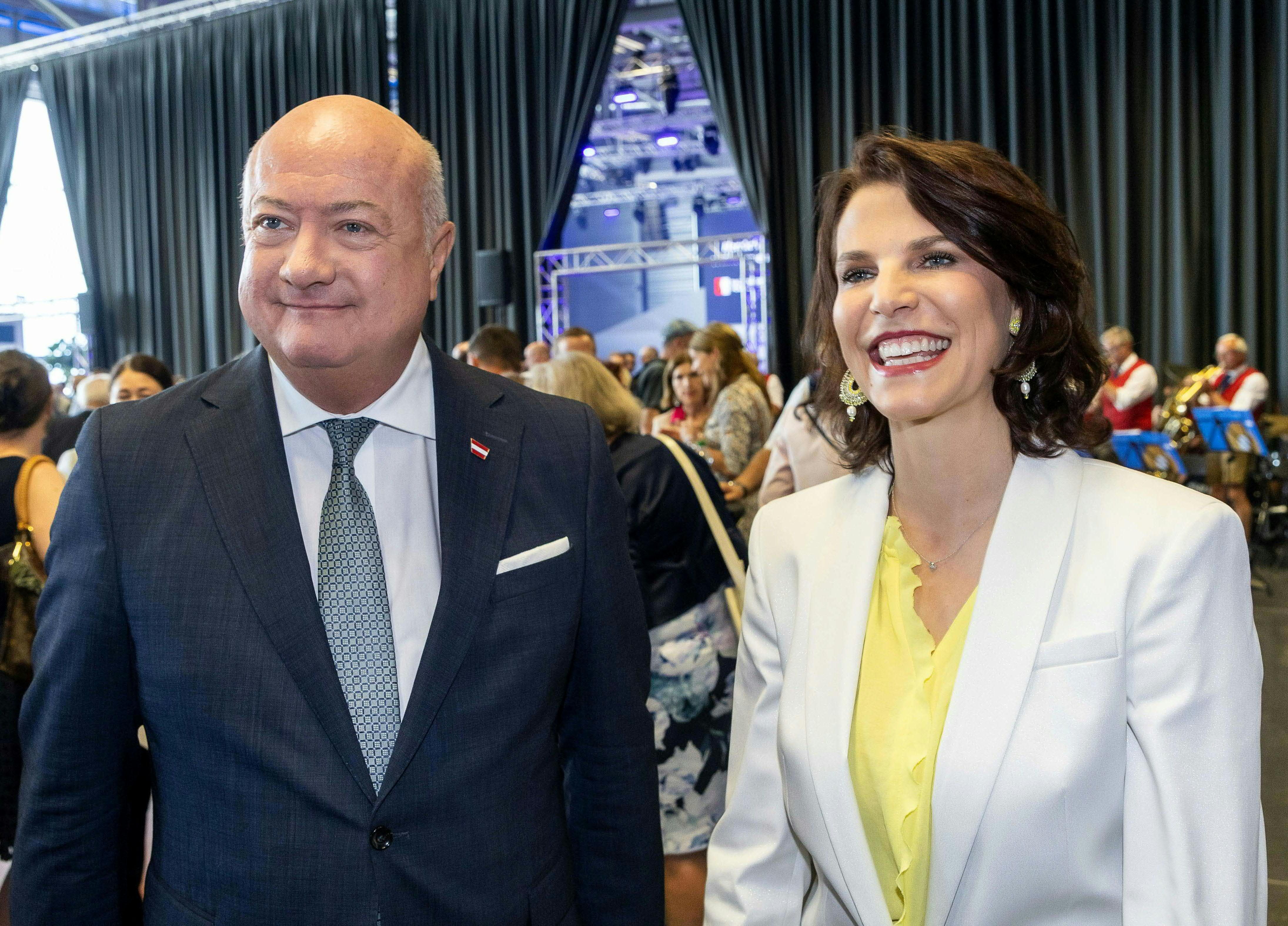 Ein Mann und eine Frau lächeln bei einer Veranstaltung mit mehreren Menschen im Hintergrund.