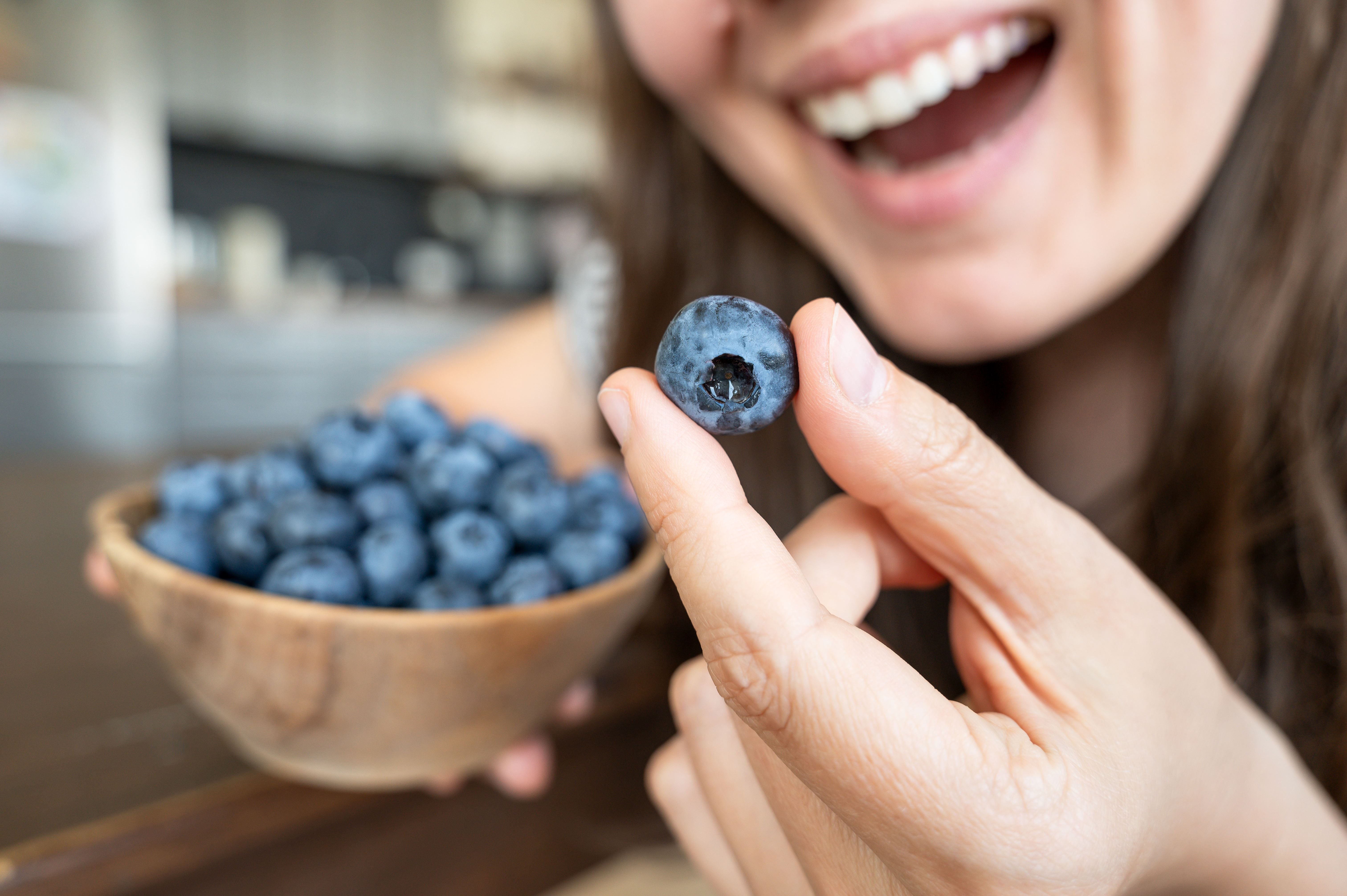 Frau hält eine Schale Blaubeeren und zeigt eine einzelne Beere lächelnd in die Kamera.