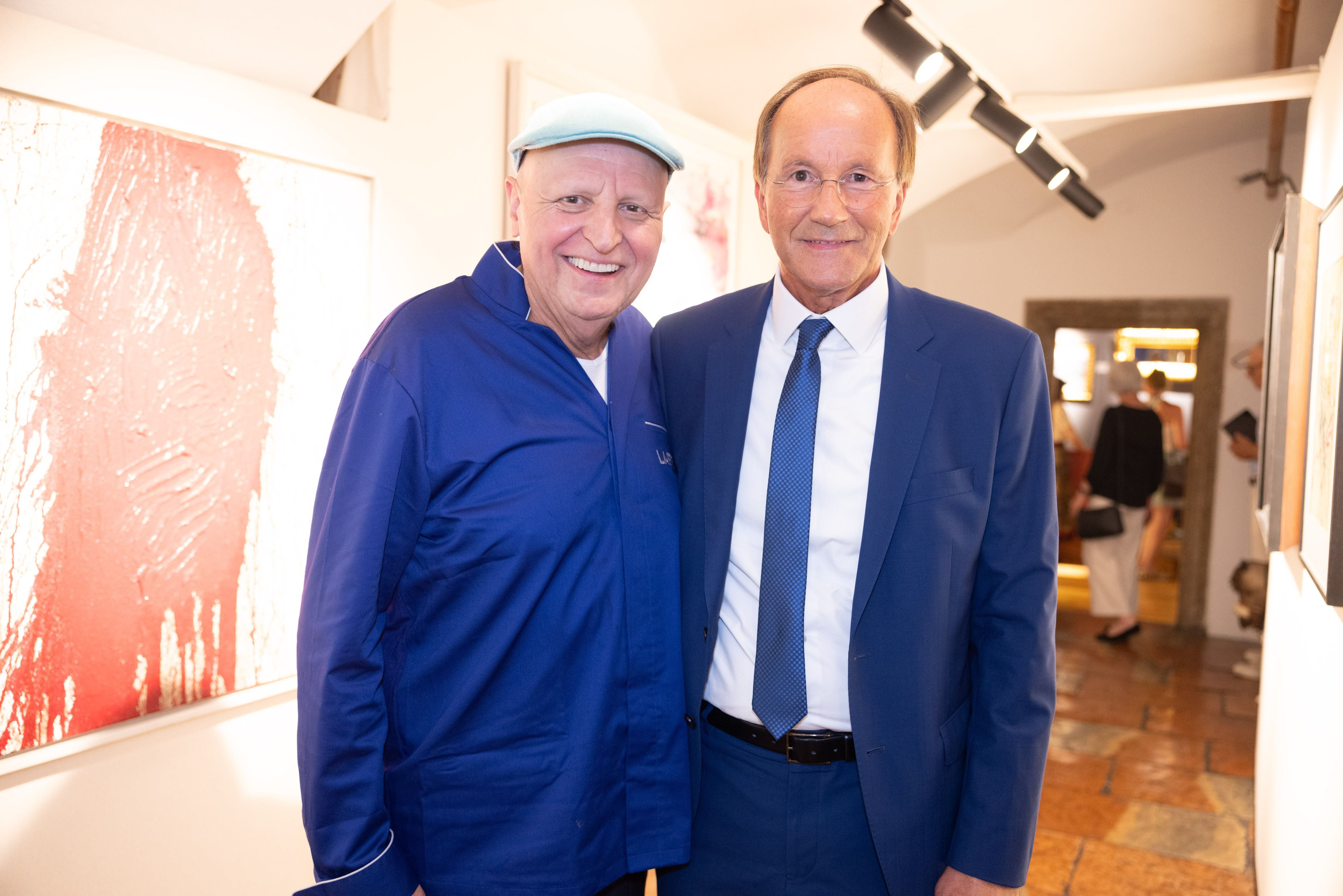 Zwei Männer in blauer Kleidung posieren lächelnd auf einer Kunstausstellung in Salzburg.