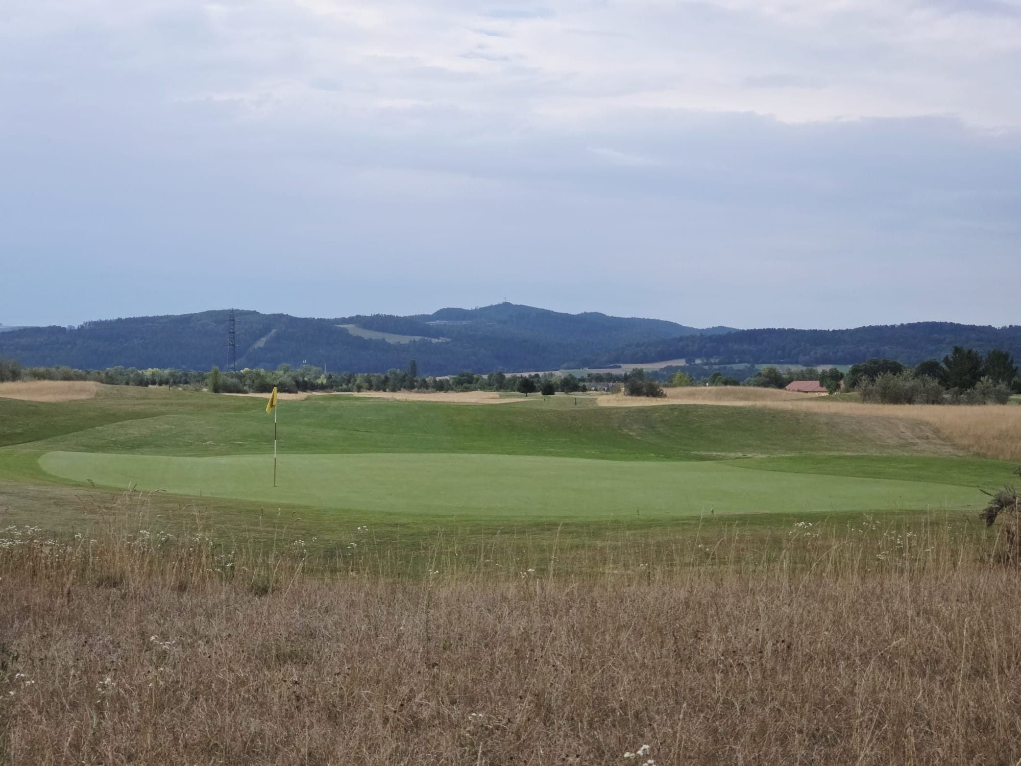 Golfplatz mit Fahne auf dem Grün, umgeben von Gras und Hügeln im Hintergrund.