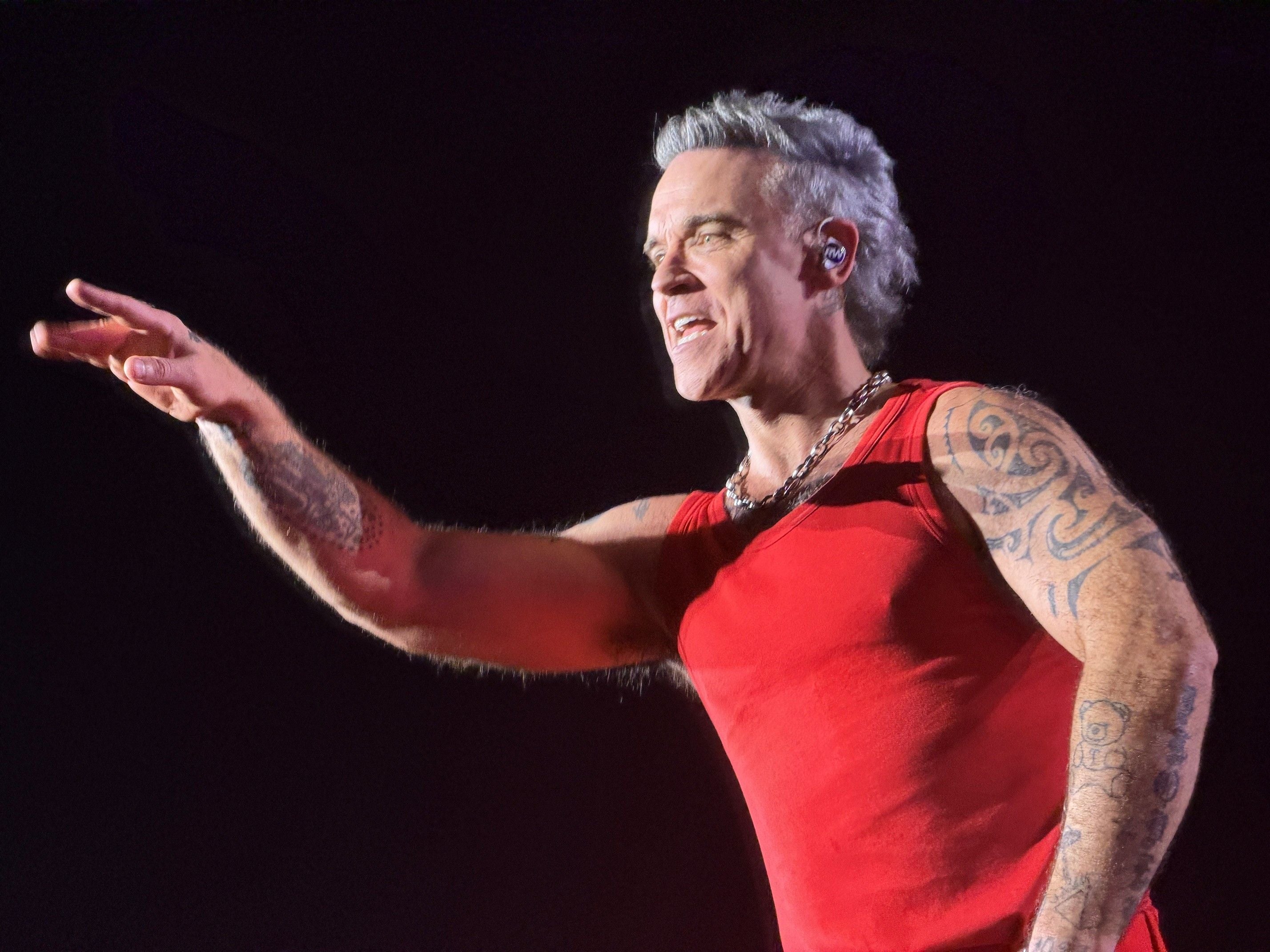 Mann mit Tattoos und rotem Shirt singt auf einer Bühne vor dunklem Hintergrund.