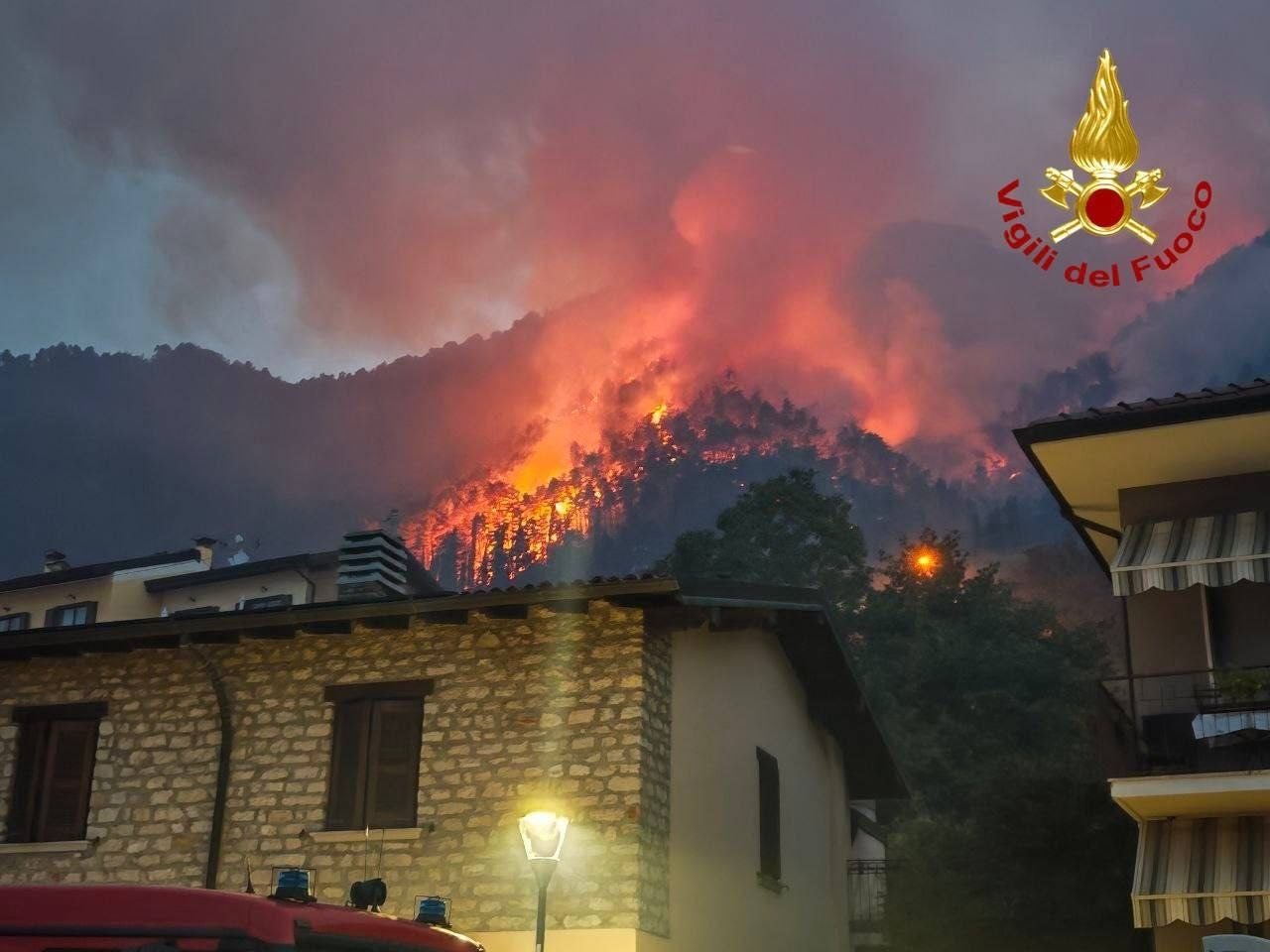 Waldbrand am Hang nahe Häusern am Gardasee, dichter Rauch und Flammen sichtbar.