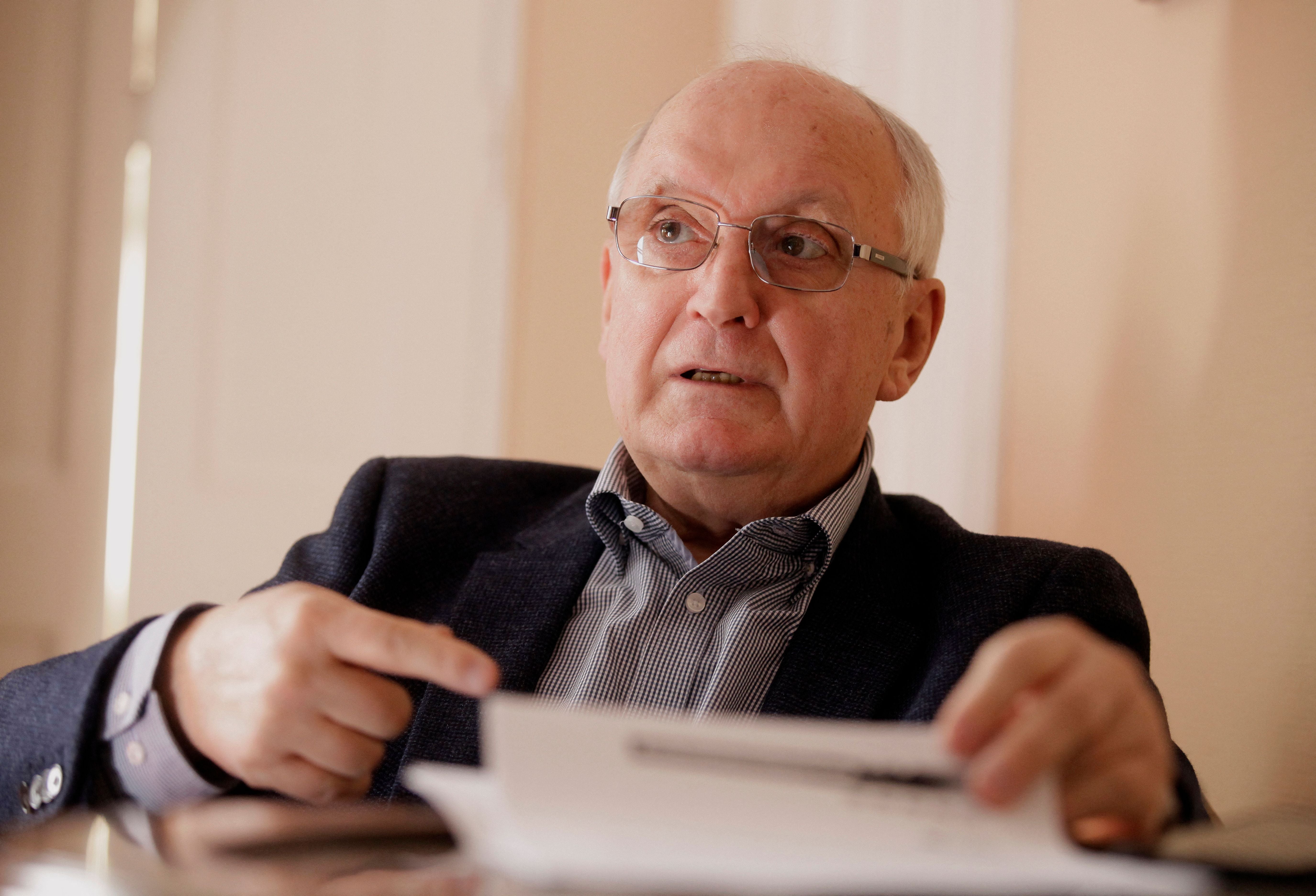 Ein älterer Mann mit Brille sitzt und spricht, während er auf ein Dokument zeigt.