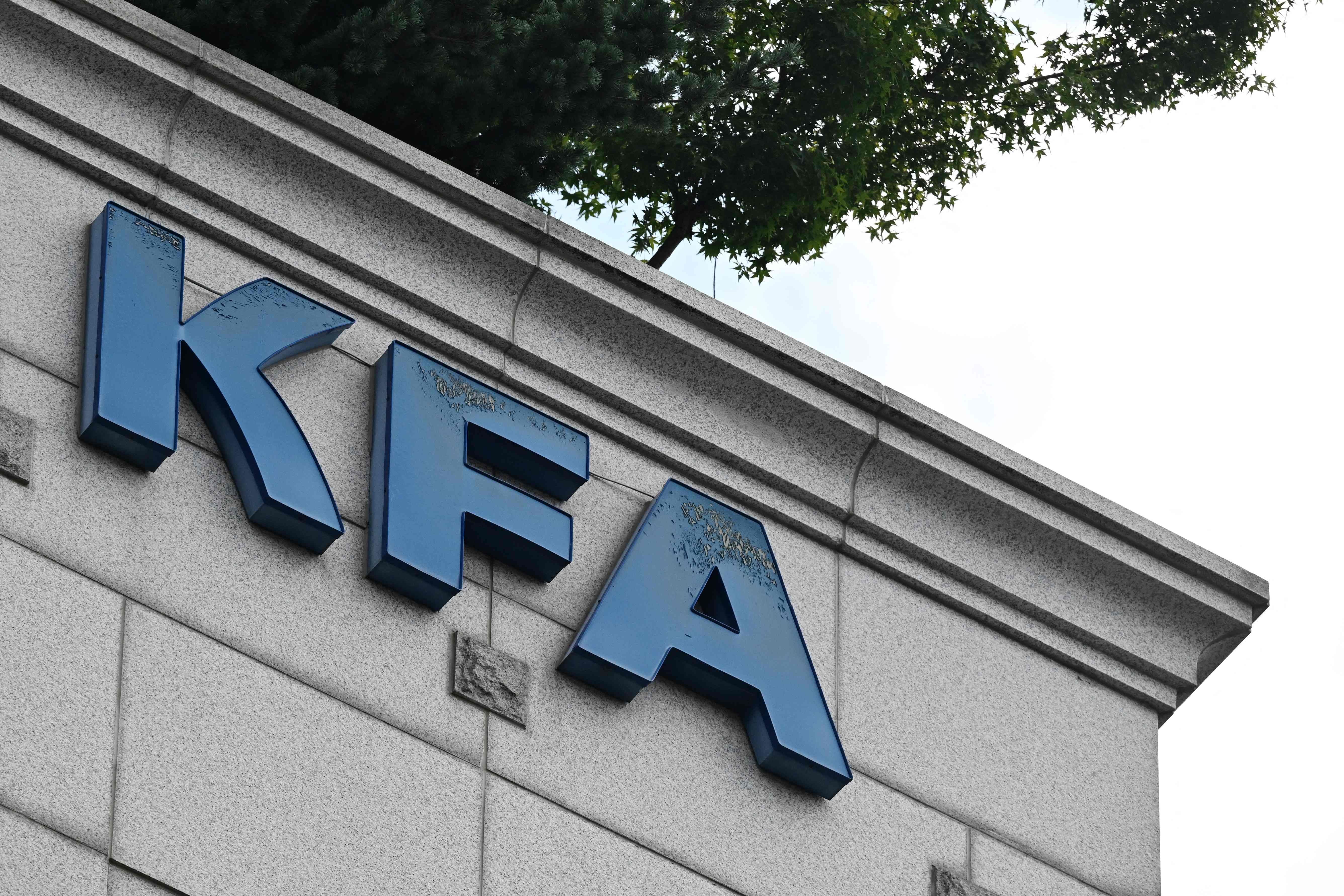 Große blaue Buchstaben KFA an einem Gebäude, darüber ein Baum und ein heller Himmel.