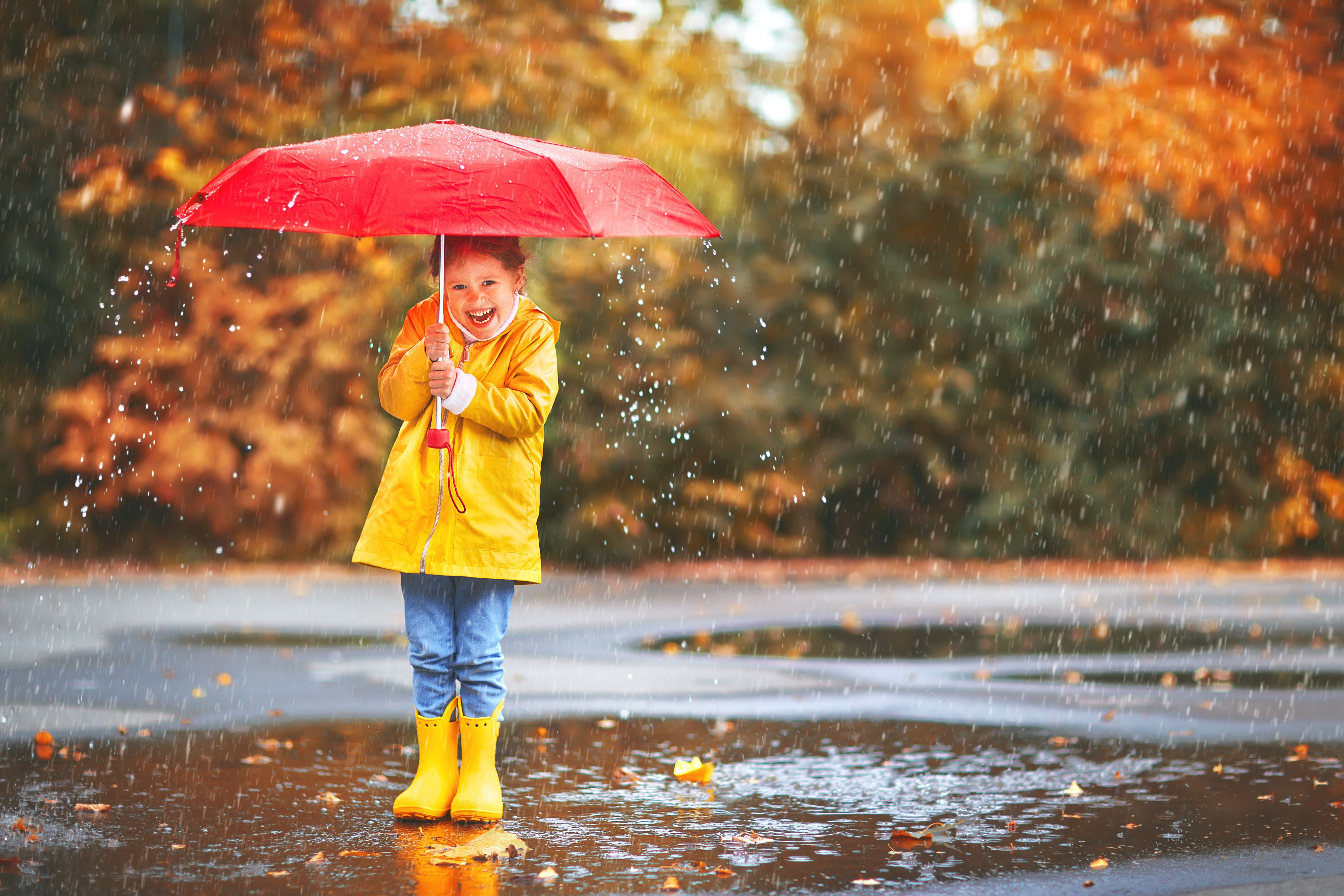 Fröhliches Kind mit rotem Regenschirm, gelber Regenjacke und Gummistiefeln im Regen.