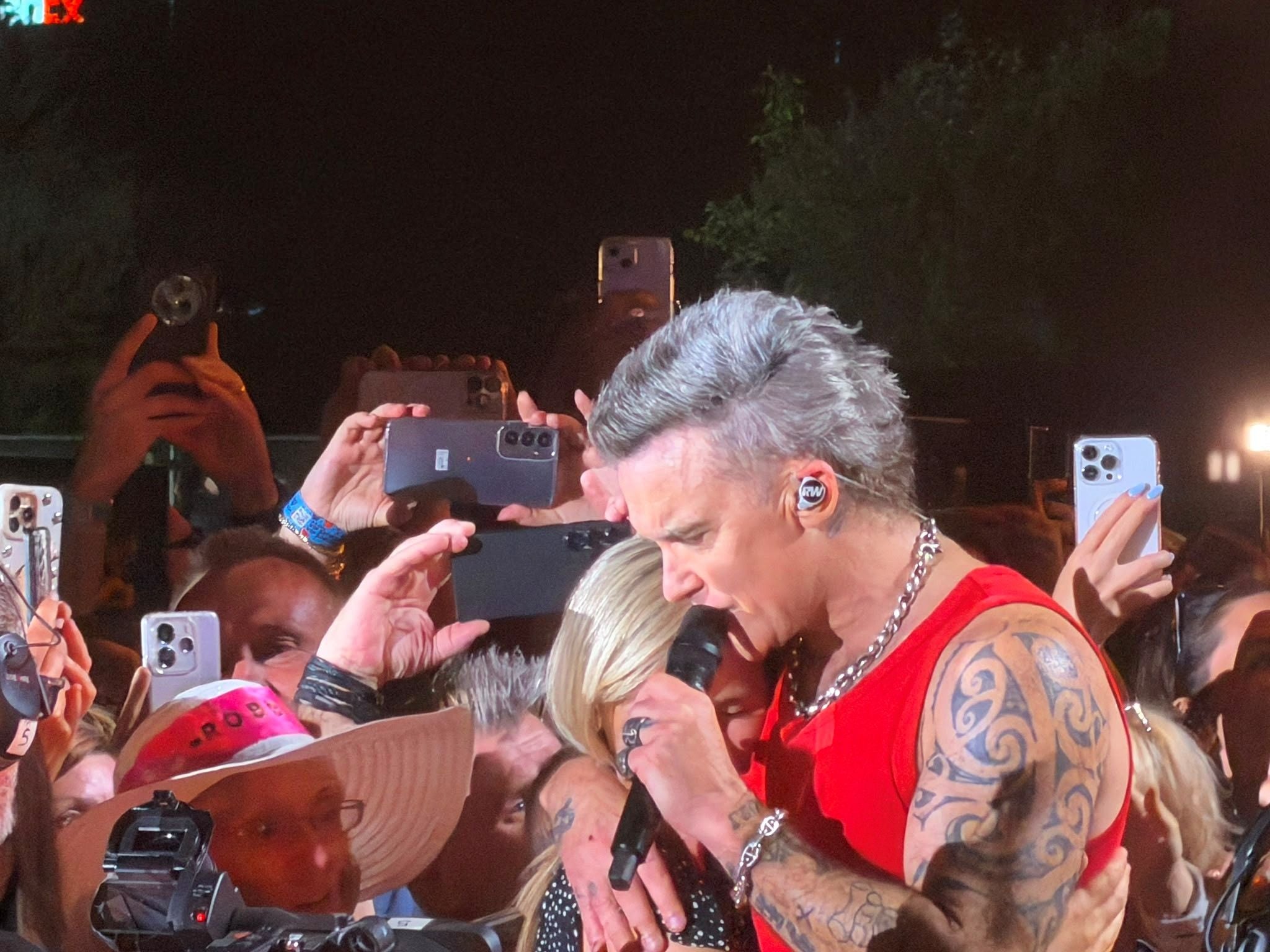 Sänger in rotem Shirt singt umringt von Fans, viele halten Handys hoch, Konzertatmosphäre.