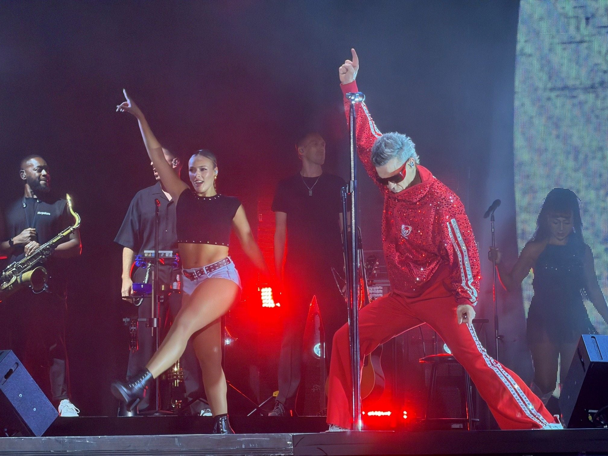 Robbie Williams und Tänzer treten auf einer hell beleuchteten Bühne auf.