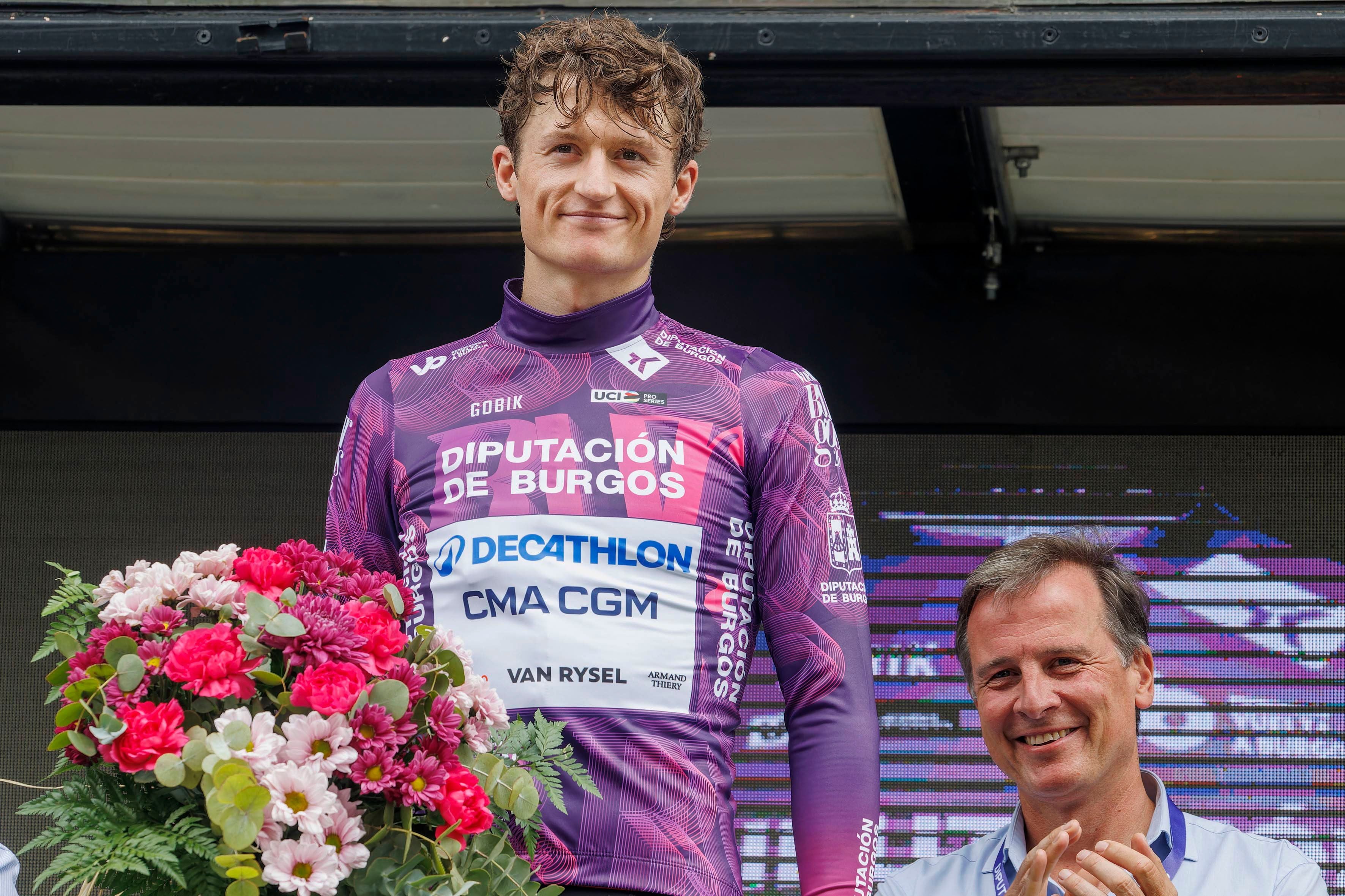 Ein Radfahrer im lila Trikot steht auf dem Podium mit Blumenstrauß, daneben ein applaudierender Mann.