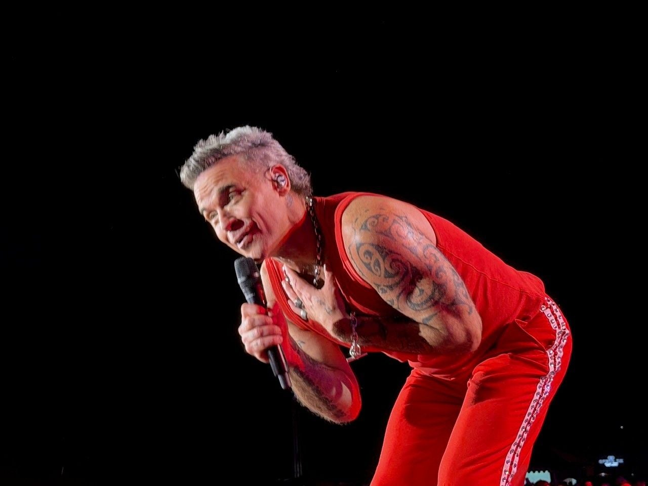 Mann in rotem Outfit mit Mikrofon und Tattoos singt auf einer Bühne vor schwarzem Hintergrund.