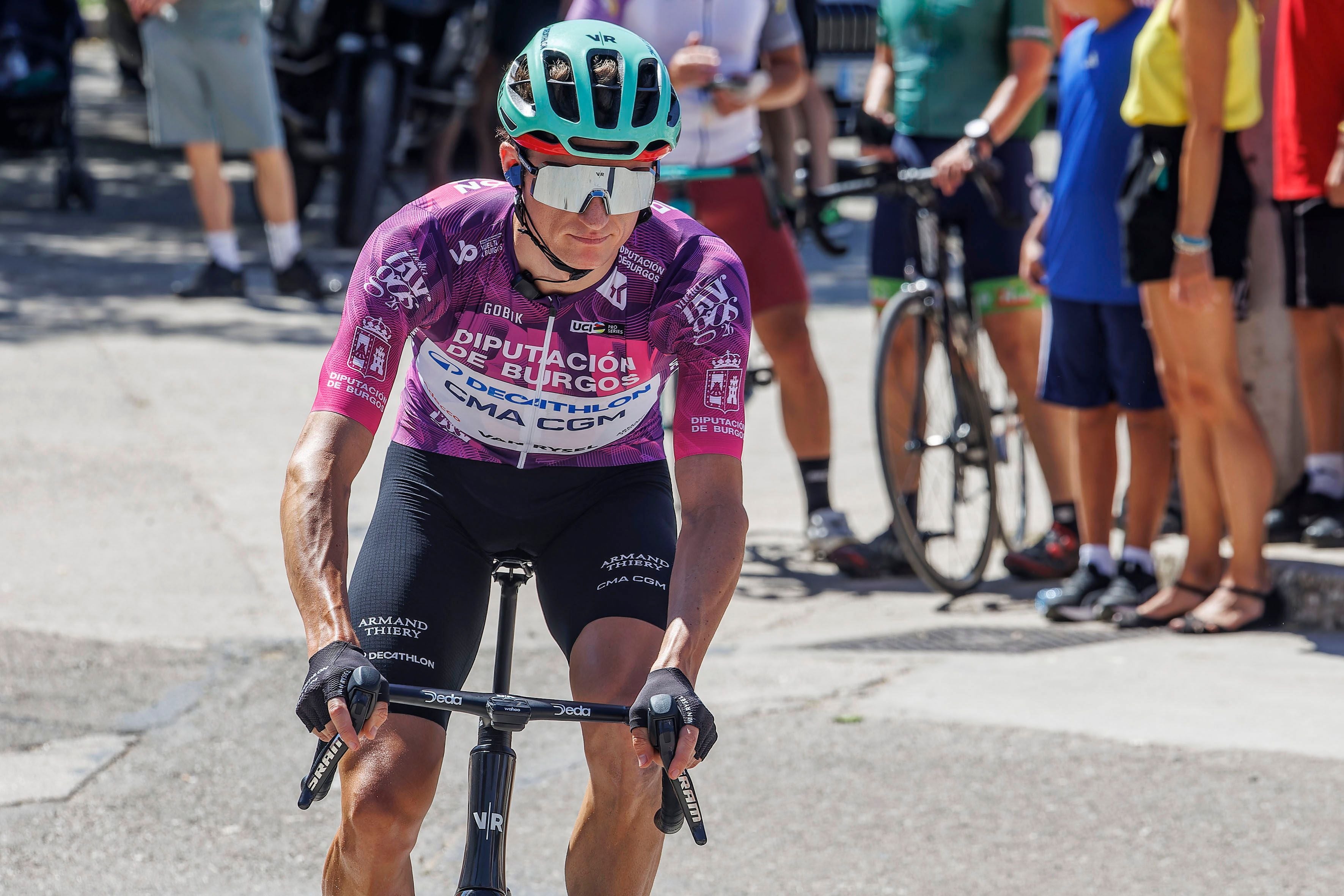 Ein Radrennfahrer im lila Trikot fährt während eines Radrennens, Zuschauer im Hintergrund.