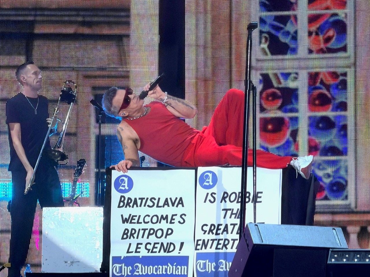Ein Sänger in rotem Outfit liegt auf einer Bühne; daneben steht ein Musiker mit Posaune.