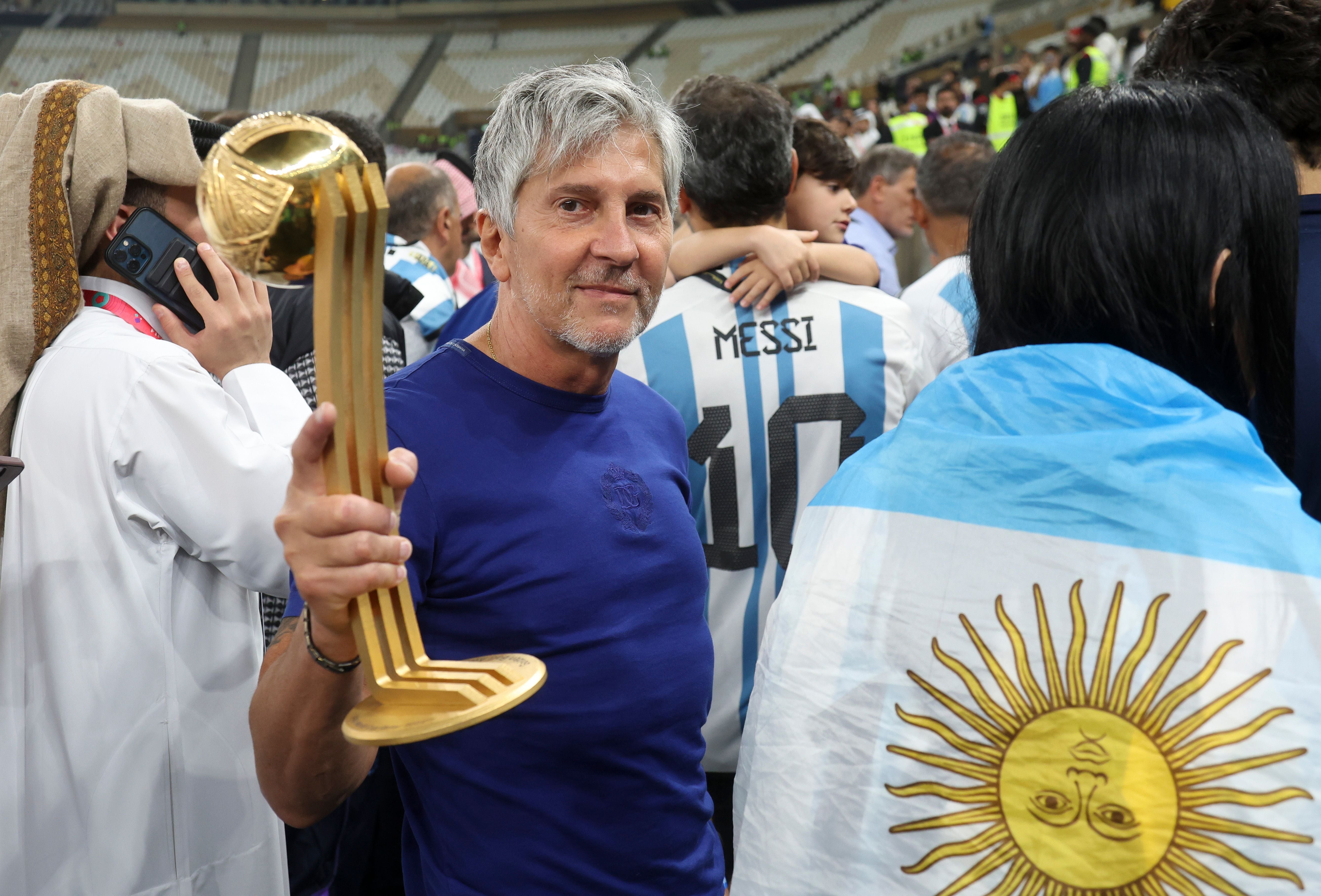Ein Mann hält eine Fußballtrophäe, umgeben von jubelnden Menschen in Argentinien-Trikots und -Flagge.