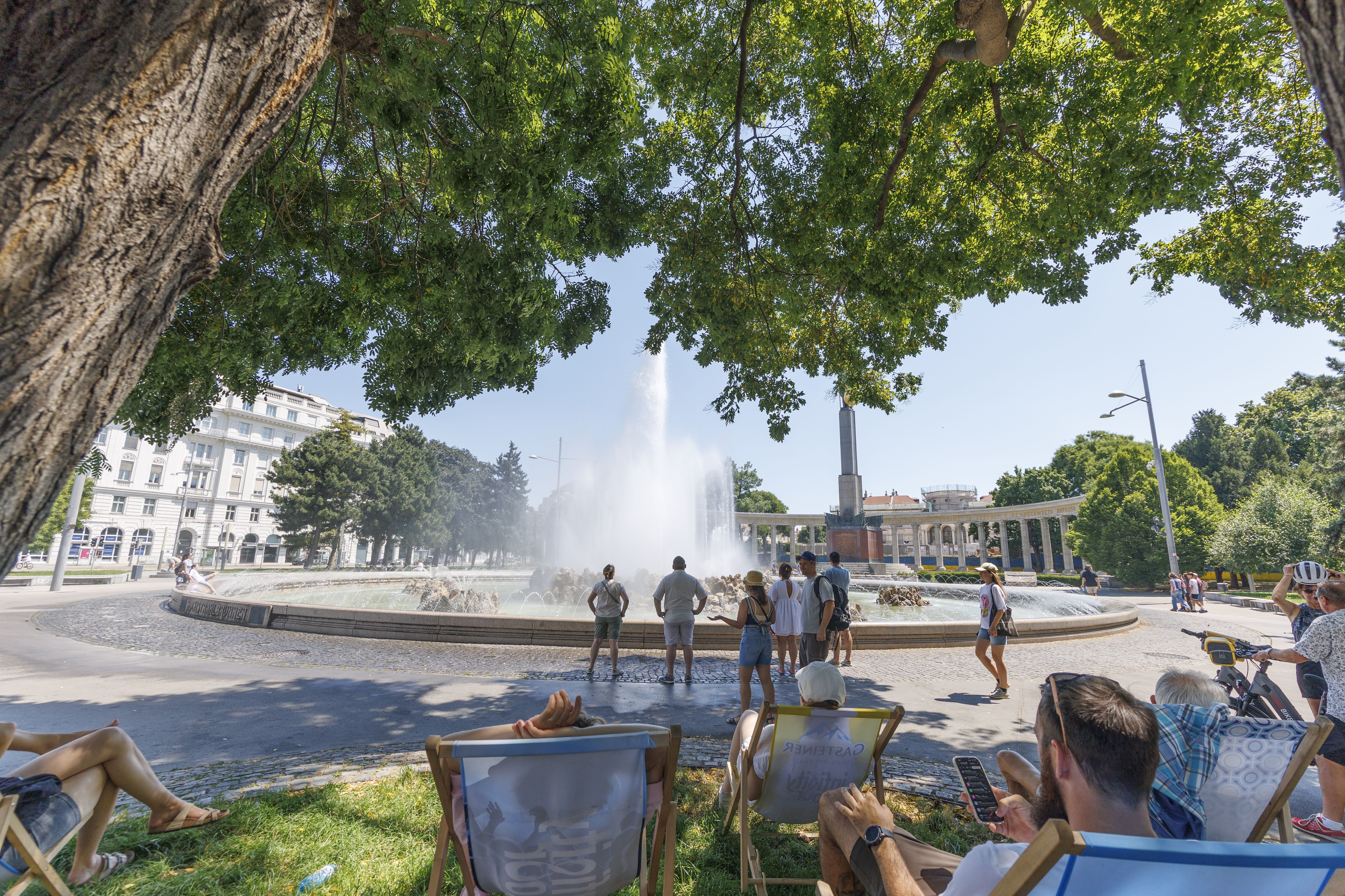Menschen suchen im Schatten vor dem Hochstrahlbrunnen in Wien Abkühlung an einem Sommertag.