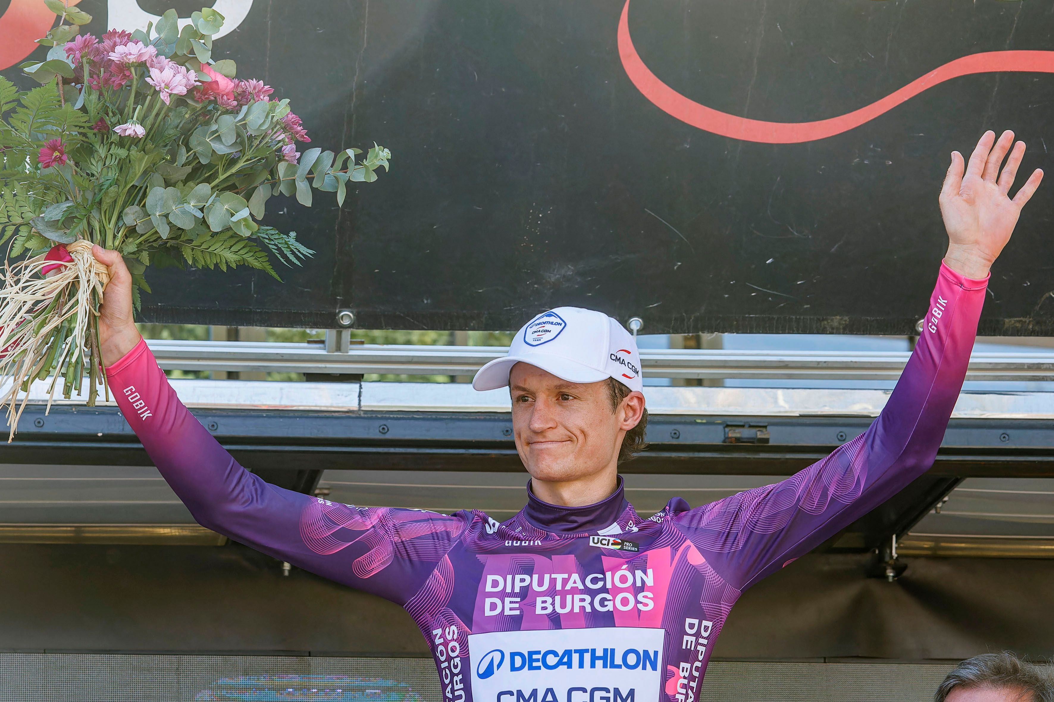 Ein Radfahrer im lila Trikot jubelt auf dem Podium und hält einen Blumenstrauß.