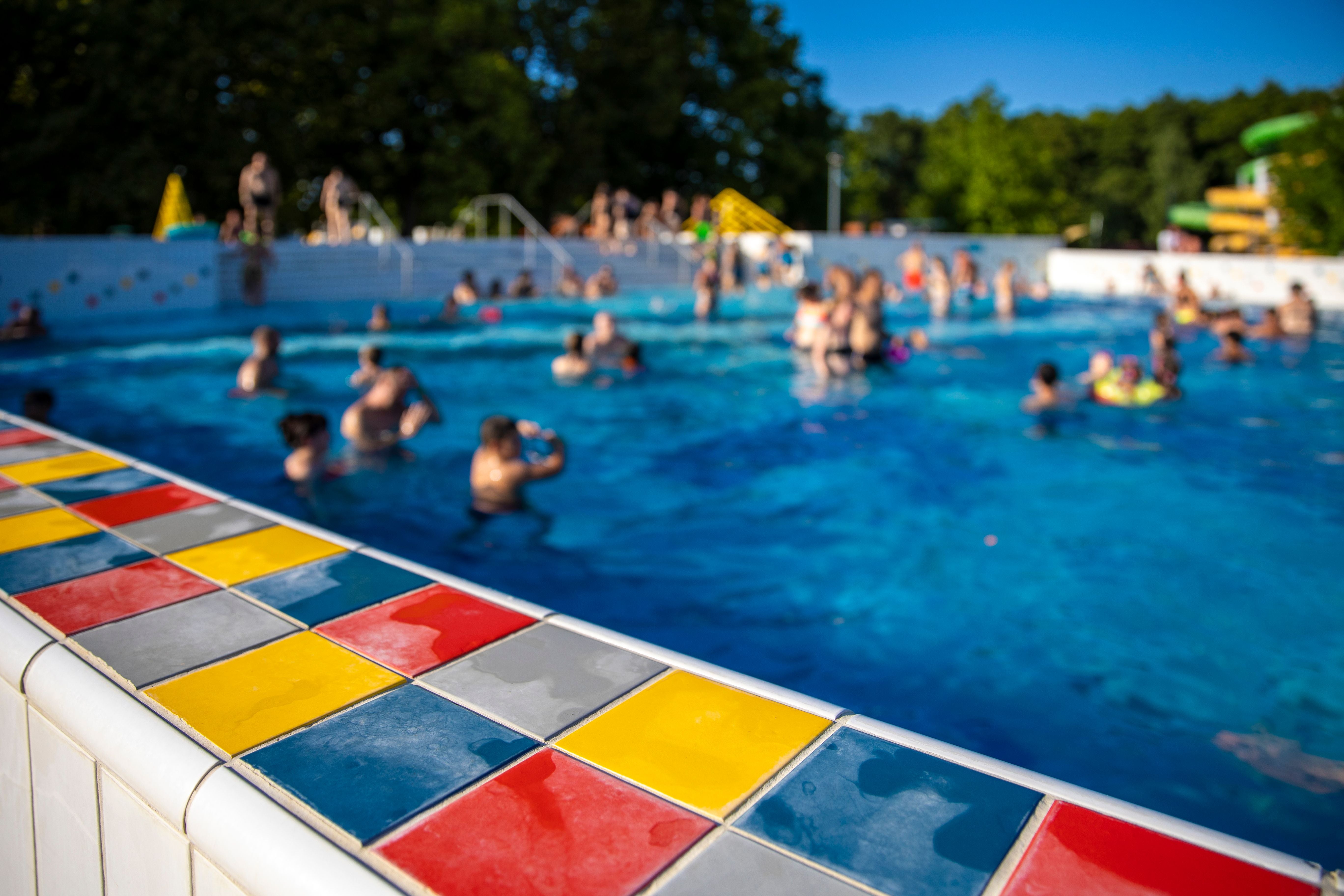 Bunt gefliester Beckenrand, viele Menschen schwimmen im Freibad bei sonnigem Wetter.