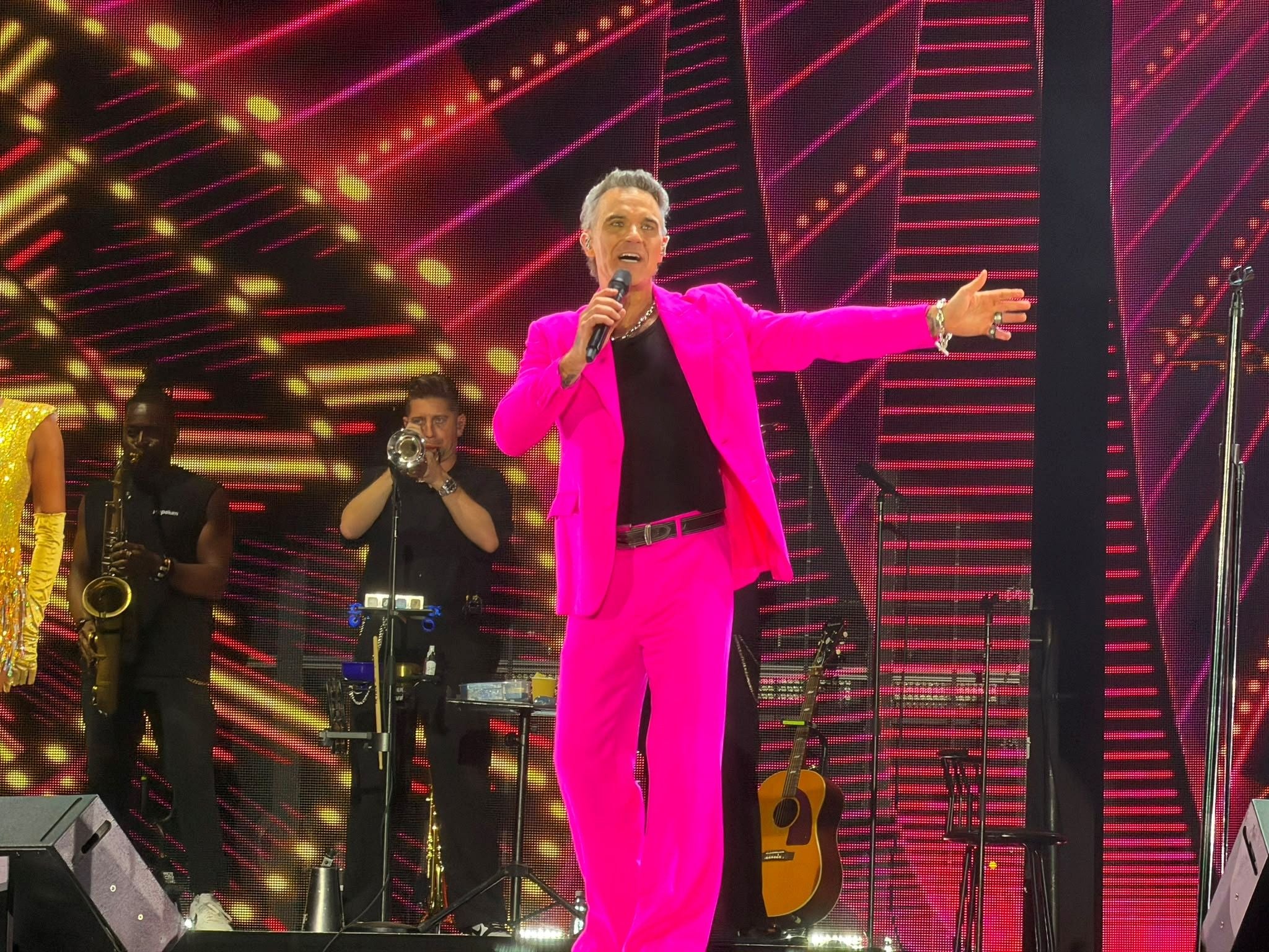 Ein Sänger im pinken Anzug performt auf einer Bühne mit Band und buntem Lichterhintergrund.