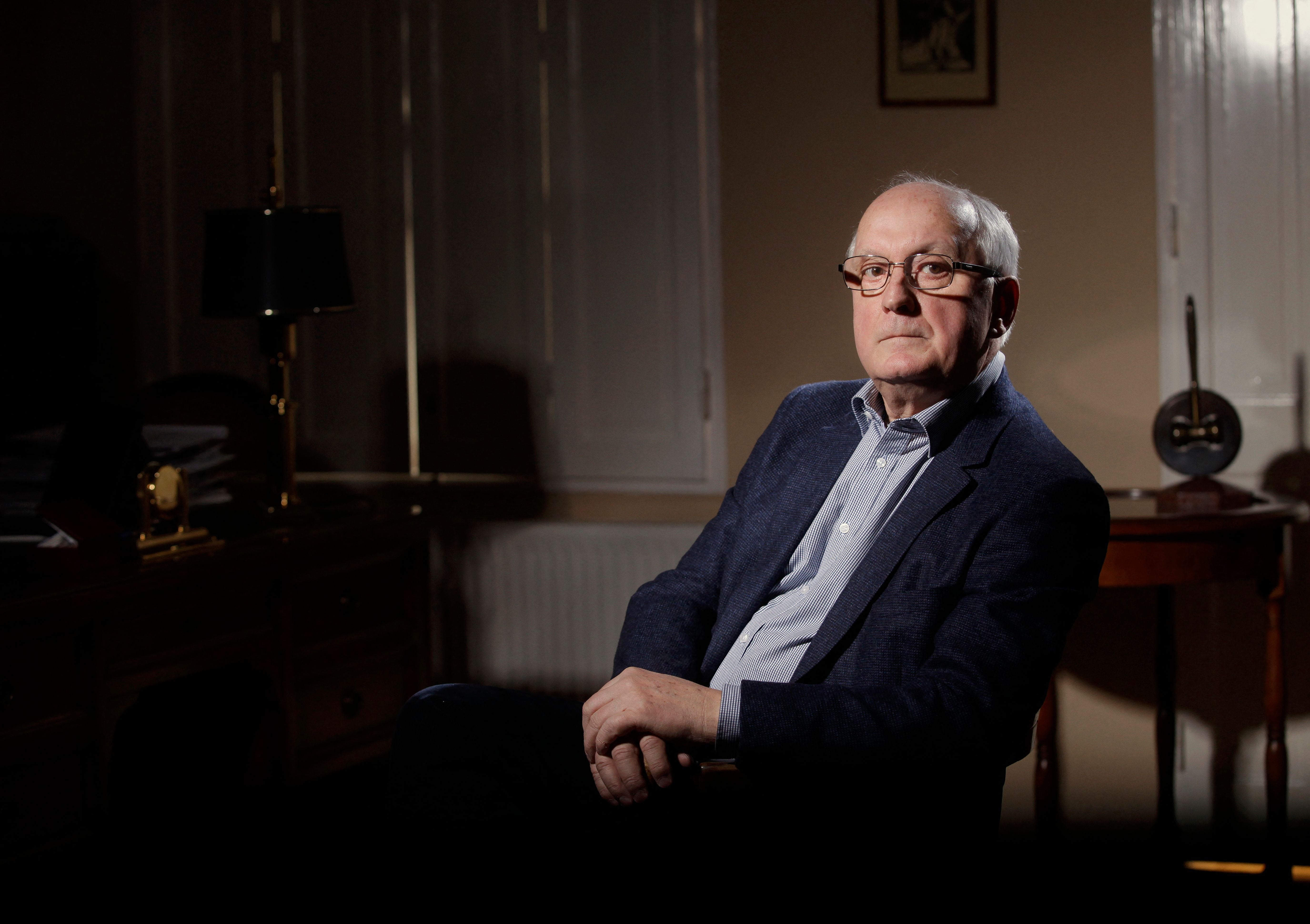 Ein älterer Mann mit Brille sitzt in einem Zimmer und blickt ernst in die Kamera.