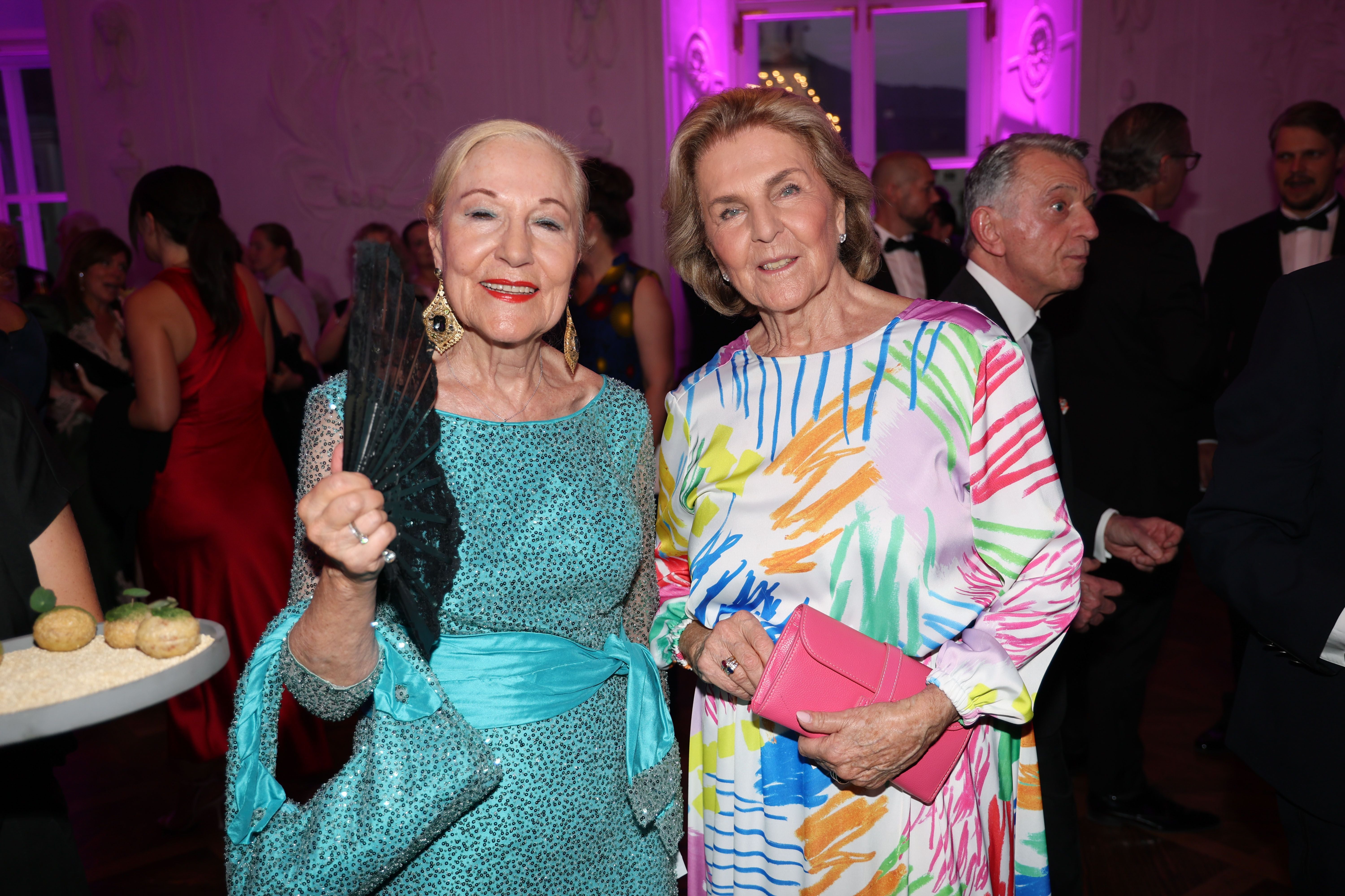 Zwei elegant gekleidete Frauen lächeln bei einer Gala mit vielen Gästen im Hintergrund.