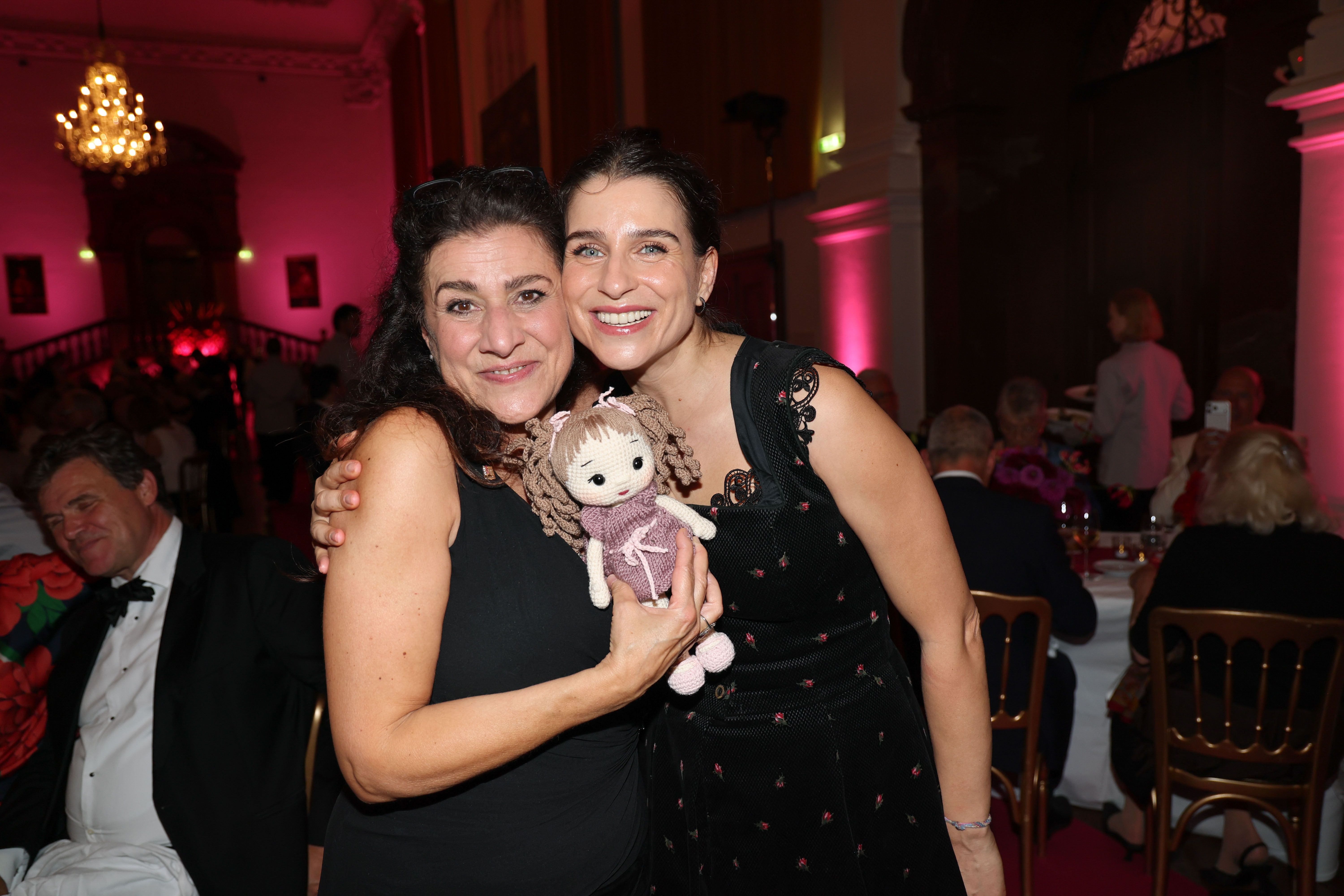 Zwei Frauen umarmen sich bei einer Gala, eine hält eine gehäkelte Puppe in der Hand.