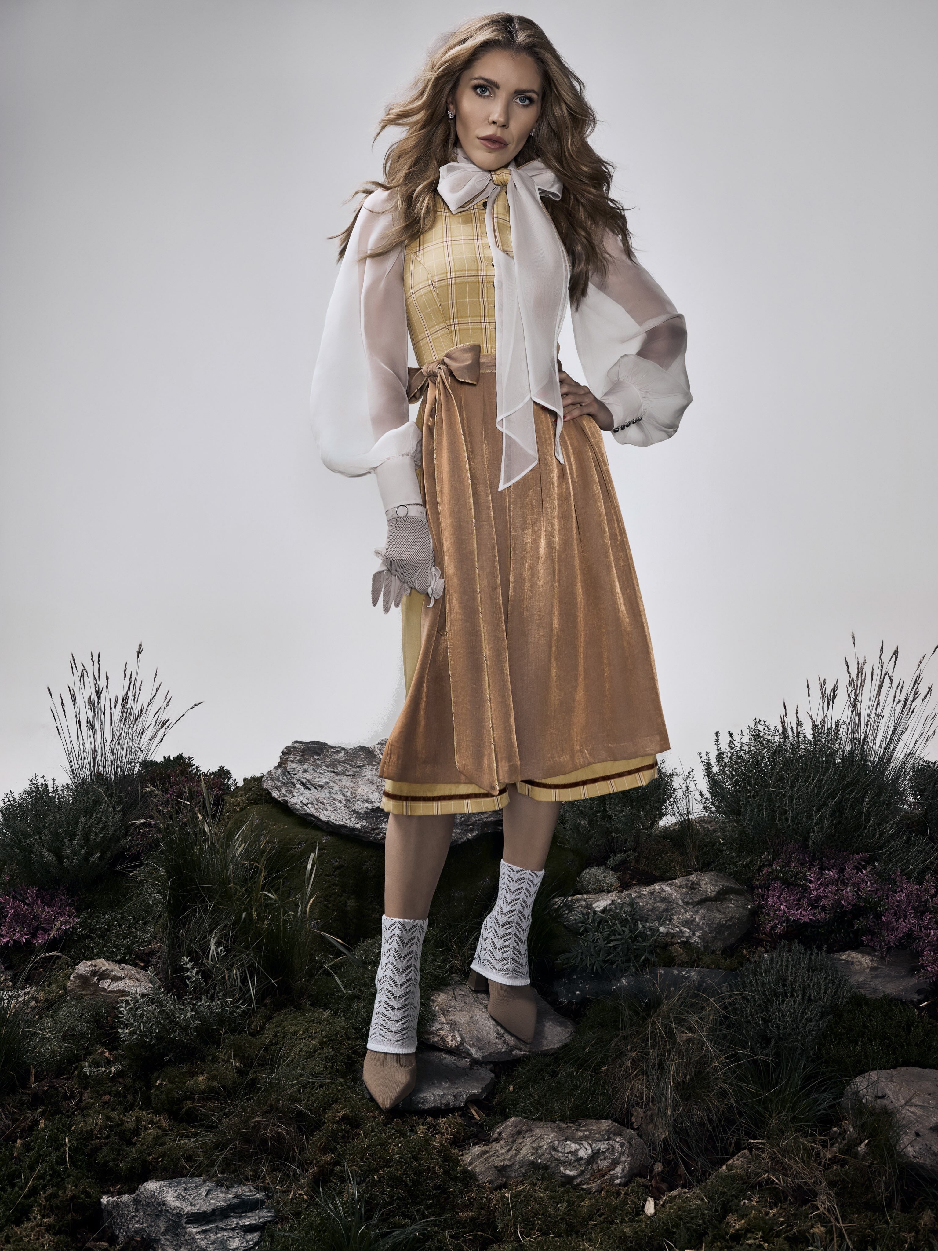 Frau im eleganten Dirndl mit Schleife steht auf moosigem Boden zwischen Steinen und Pflanzen.