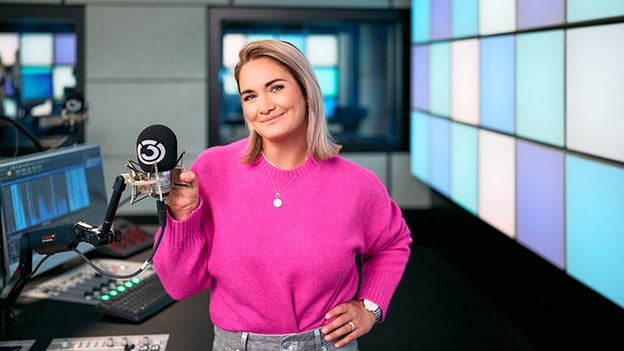 Frau mit pinkem Pullover steht lächelnd in einem modernen Radiostudio vor einem Mikrofon.