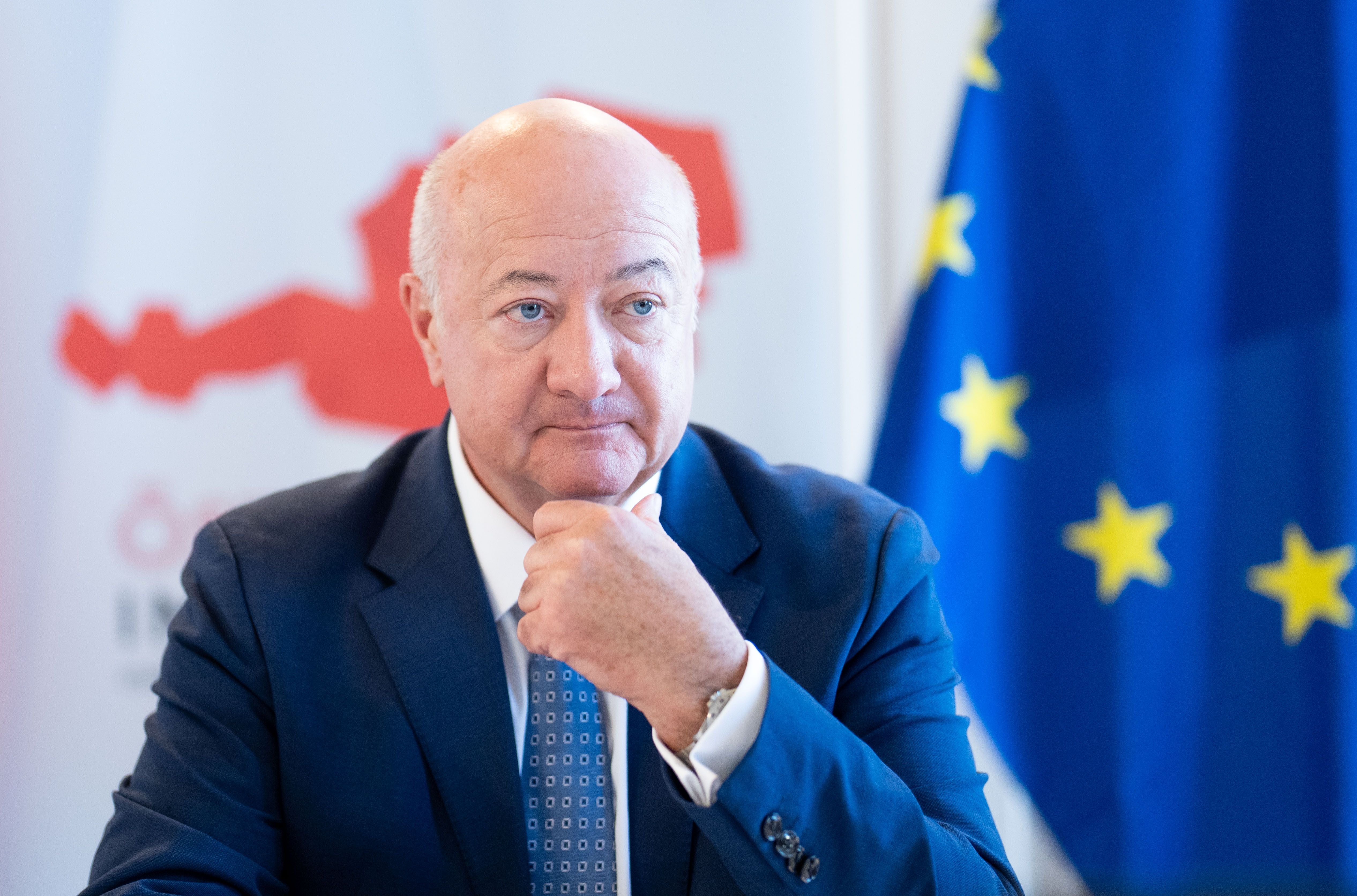 Ein Mann im Anzug sitzt nachdenklich vor einer EU-Flagge und einem Österreich-Logo.