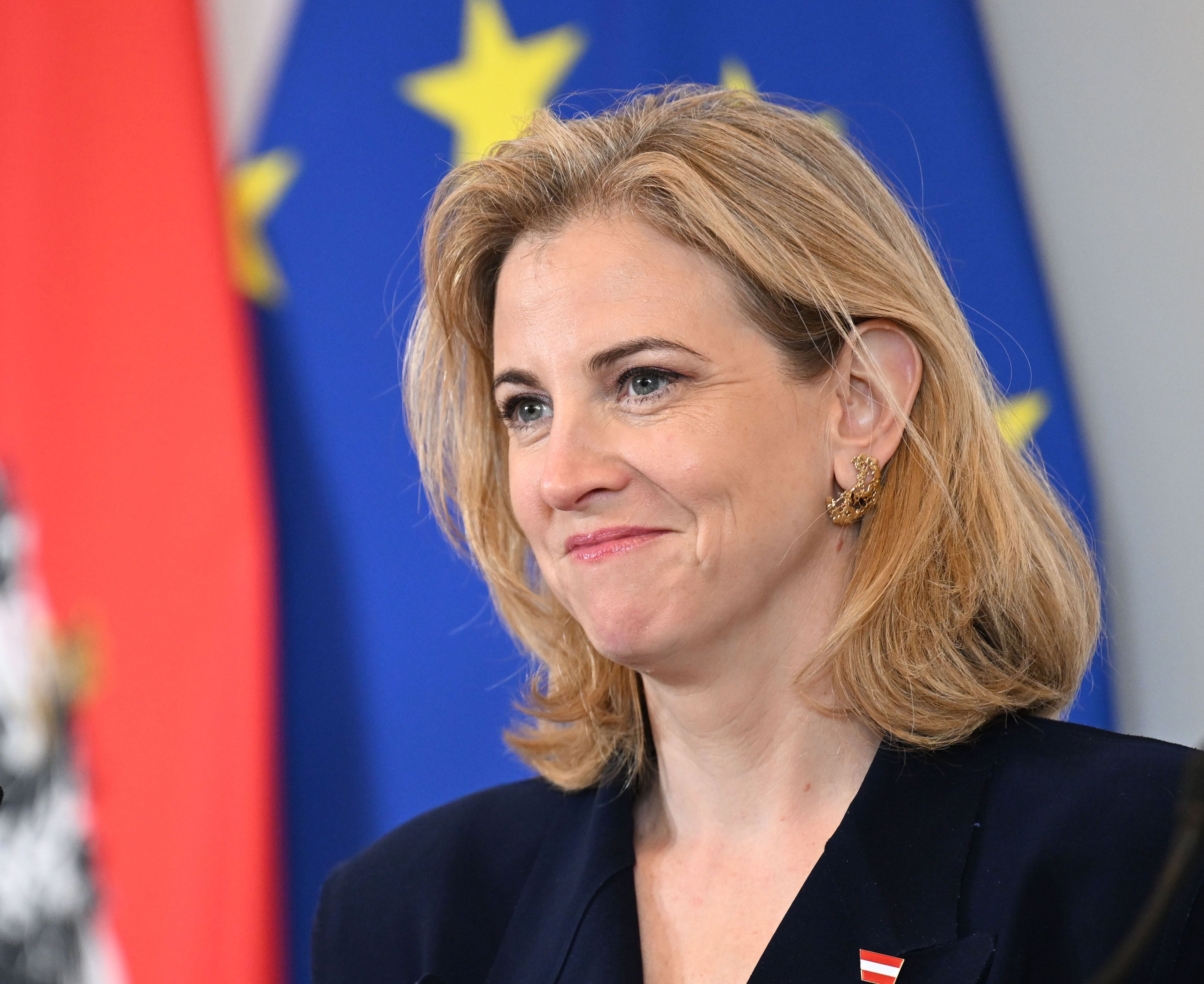 Eine Frau mit blondem Haar steht vor EU- und Österreich-Flaggen und lächelt leicht.