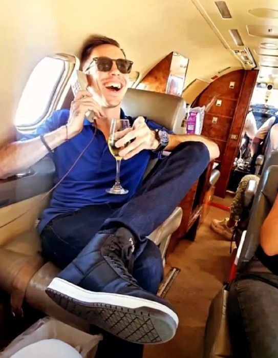 Mann mit Sonnenbrille telefoniert und trinkt Wein im Privatjet, wirkt entspannt und lachend.