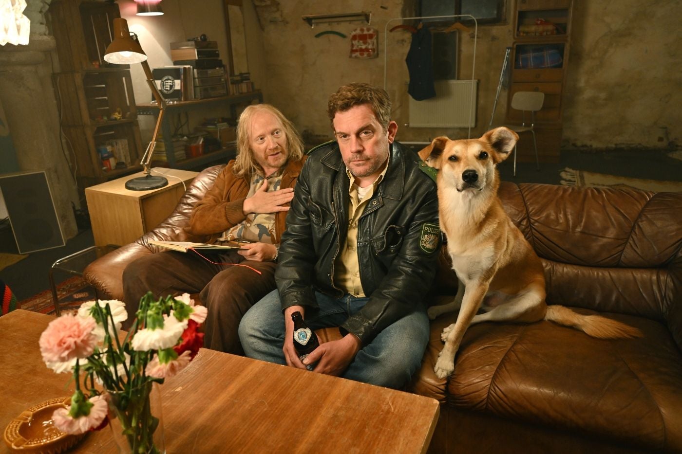 Zwei Männer und ein Hund sitzen auf einem braunen Ledersofa in einem gemütlichen Wohnzimmer.