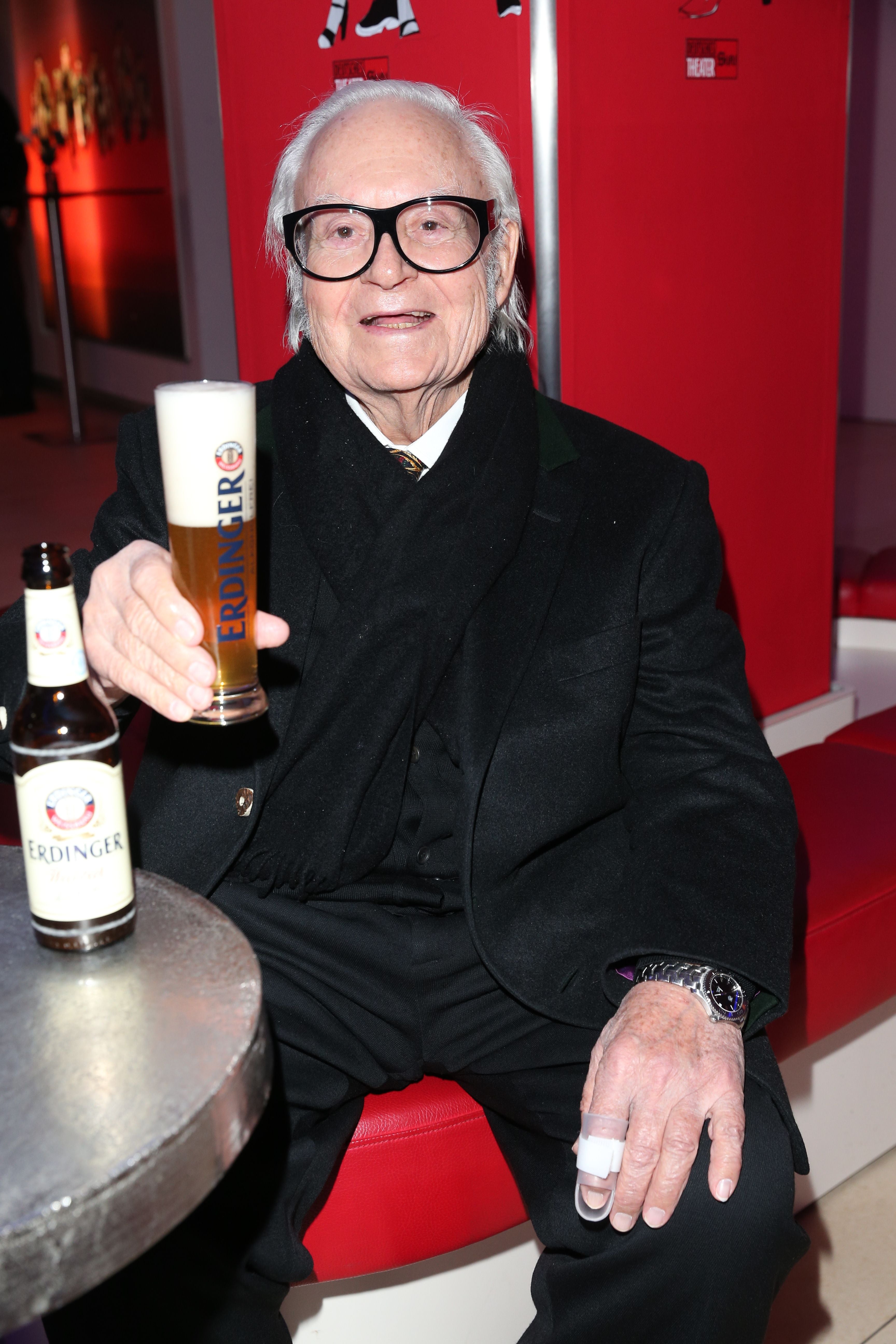 Älterer Mann im Anzug hält ein Glas Erdinger Weißbier und lächelt in einer roten Sitzbank.